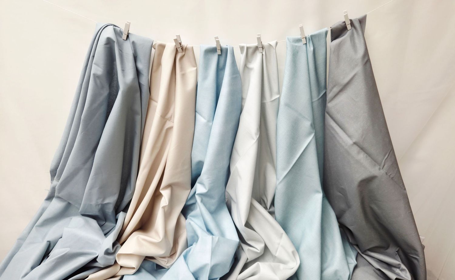 Multiple fabric swatches in soft blue, gray, beige, and white tones hanging from a rod against a light background.