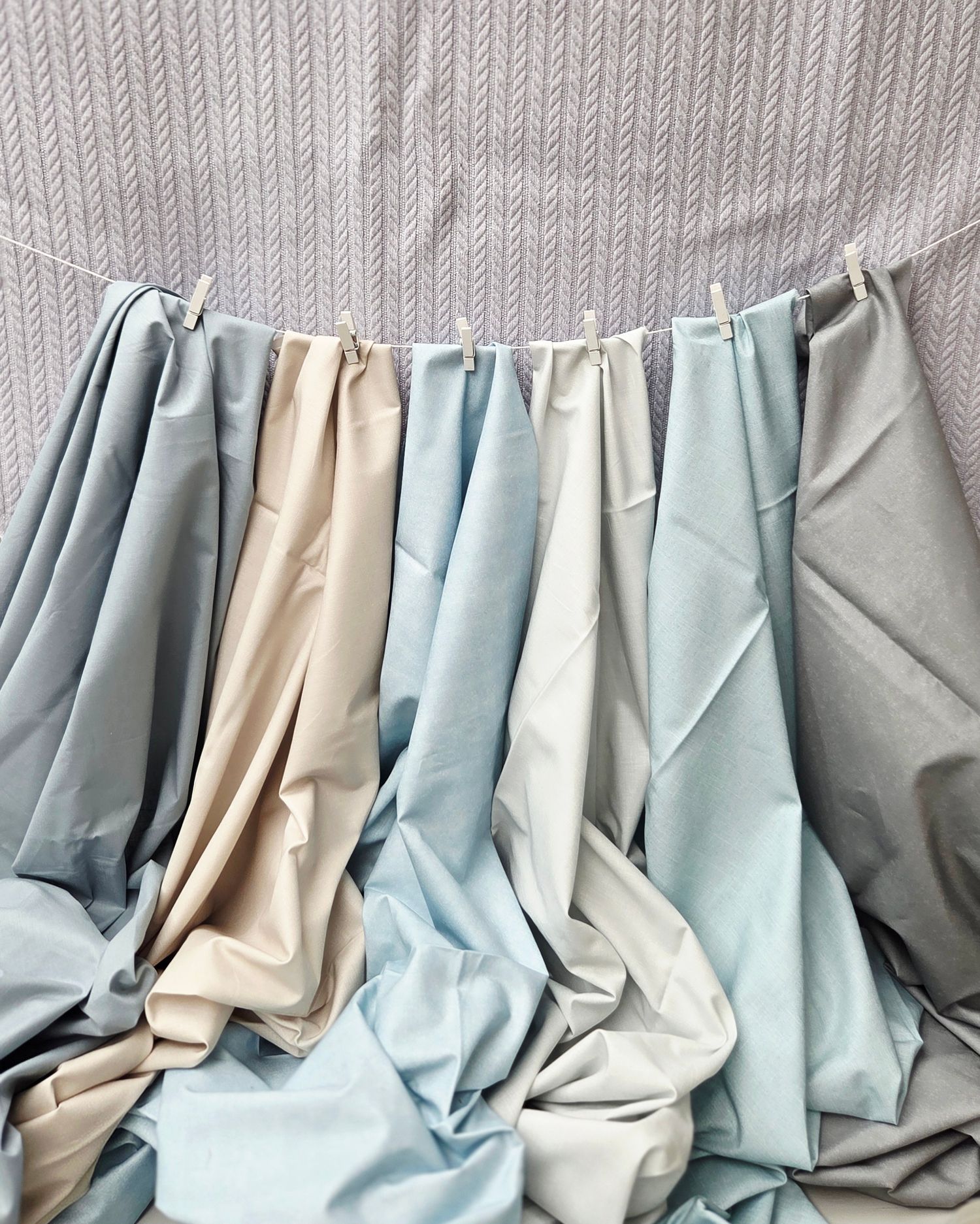 Soft pastel fabric swatches in blue, gray, and beige tones draped elegantly on a clothesline against a herringbone wall.