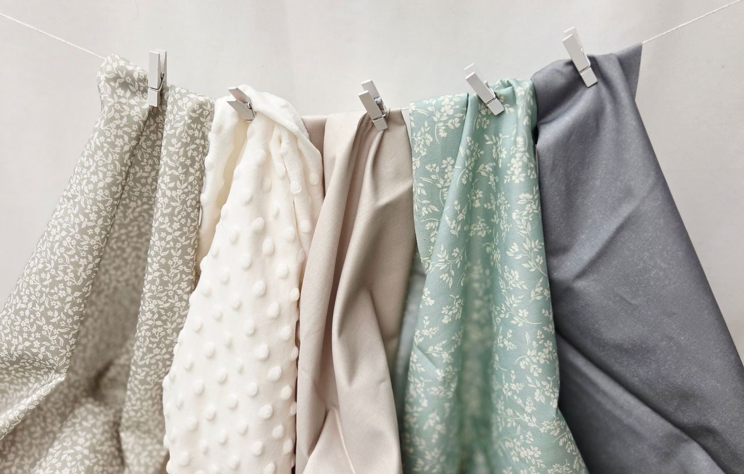 Five soft fabric swatches in neutral tones hang on a line, including white, beige, mint, and gray textiles.