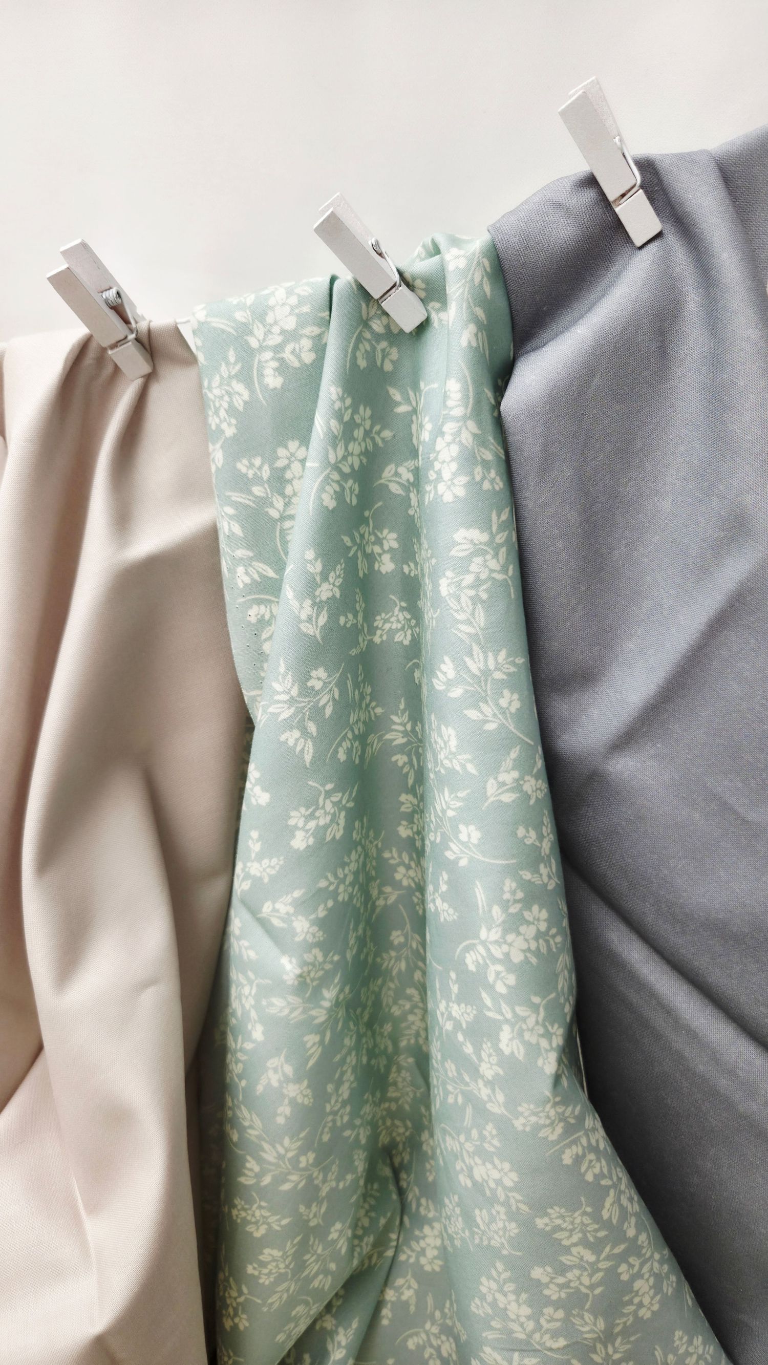 Three fabric swatches in beige, mint floral, and gray hang from white clothespins against a white background.