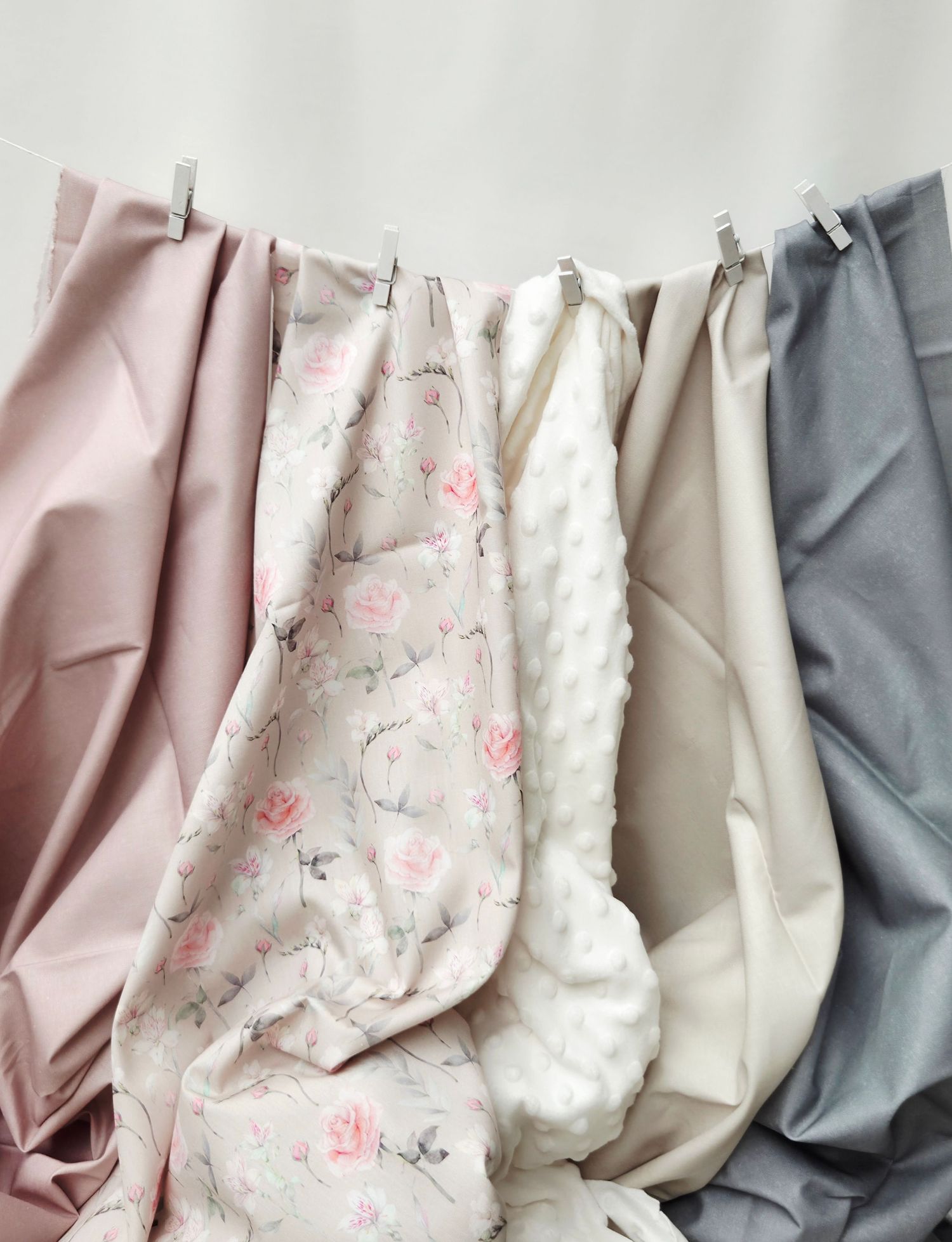 Four soft baby blankets in pink, floral, cream, and gray hang from a clothesline with clips against a white wall.