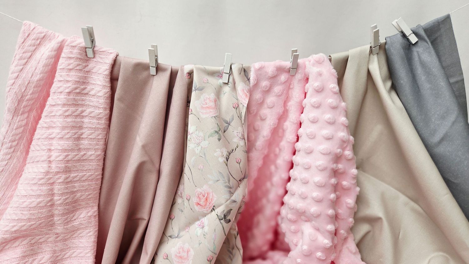 Assorted pink and gray baby blankets hanging on a clothesline with wooden clips, showcasing various textures and patterns.
