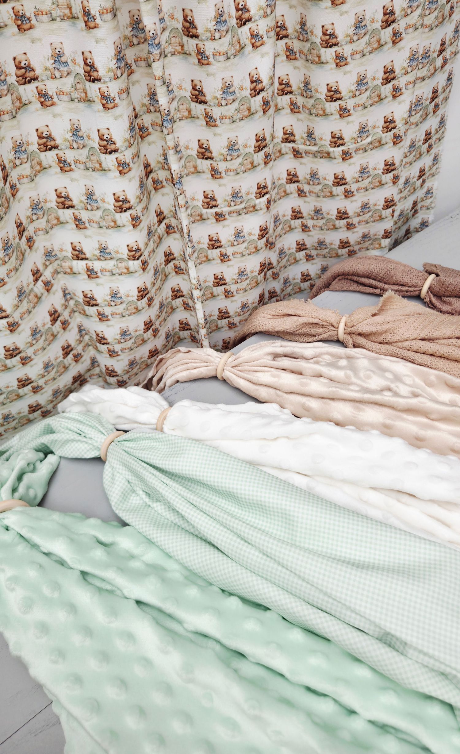 Assorted baby blankets in mint green, white, and blush pink stacked near teddy bear print curtains.