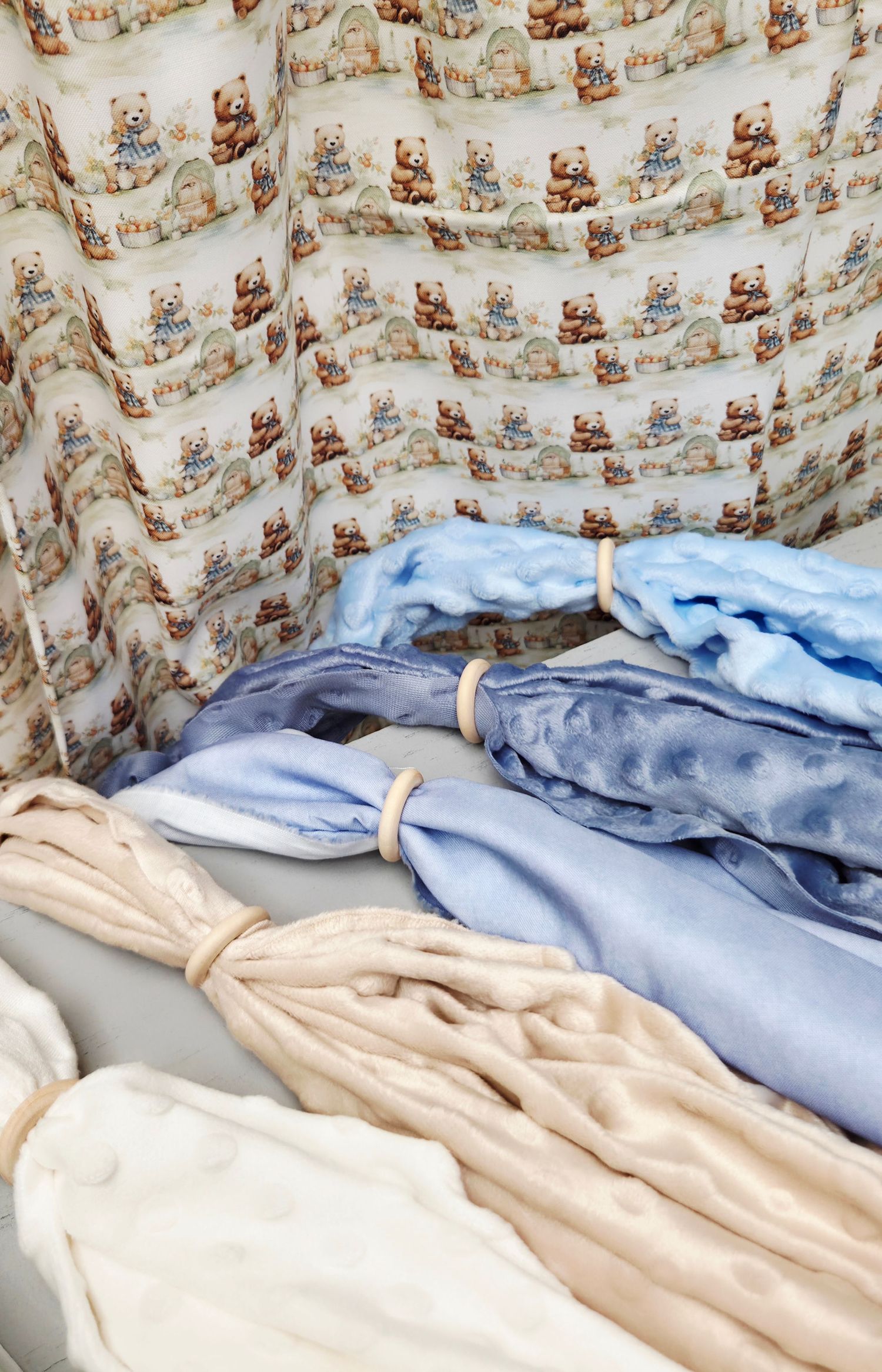 Rolled baby blankets in blue, beige, and white beside a teddy bear print fabric backdrop.