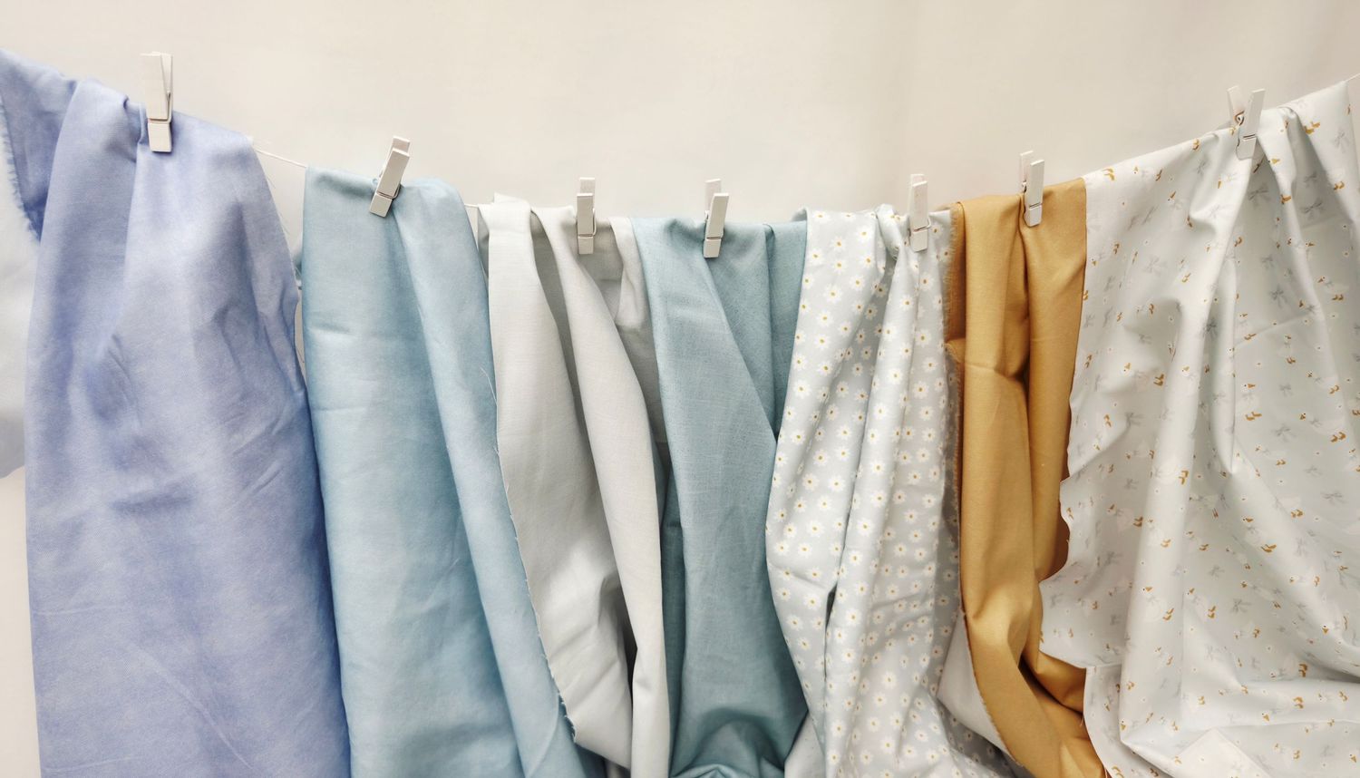 Colorful fabric swatches in blue, white, and gold hanging on a clothesline with white clips against a white wall.