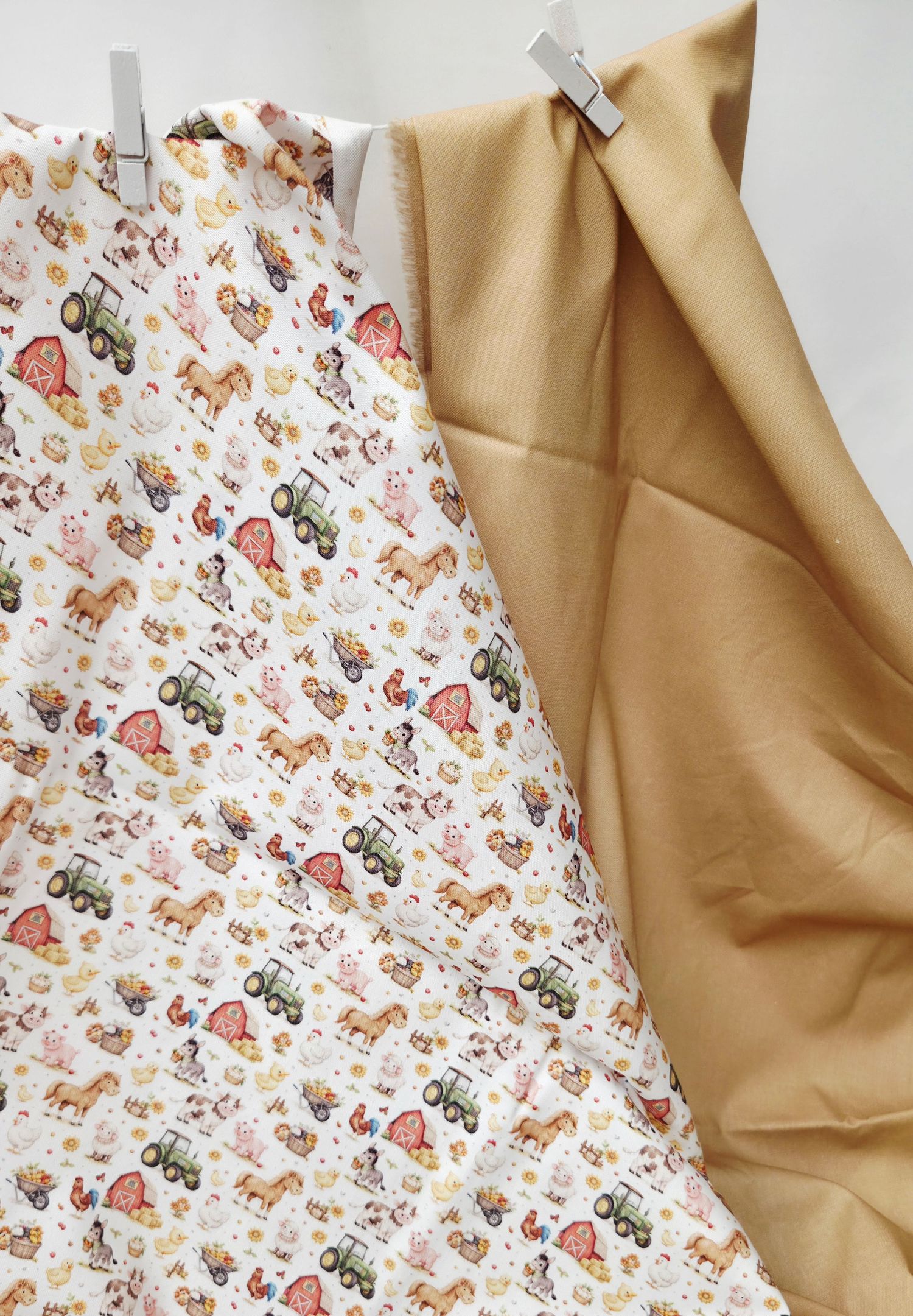 Two fabric swatches hanging on clips, one with farm animal print and one solid tan cotton fabric.