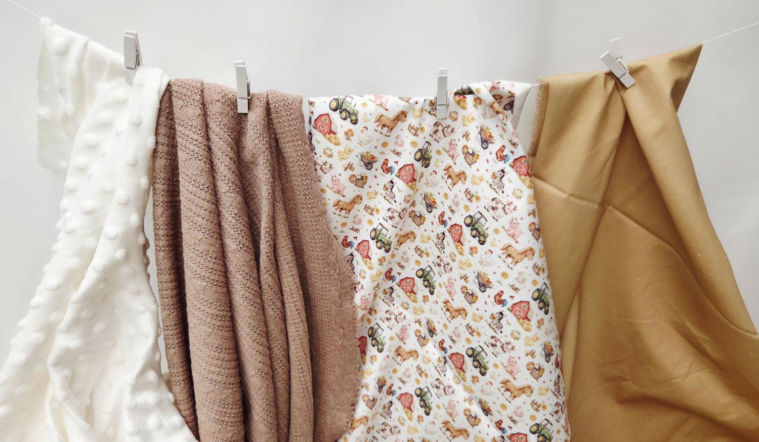 Four decorative fabrics hanging on a clothesline, featuring white, dusty rose, floral print, and golden tan textiles.