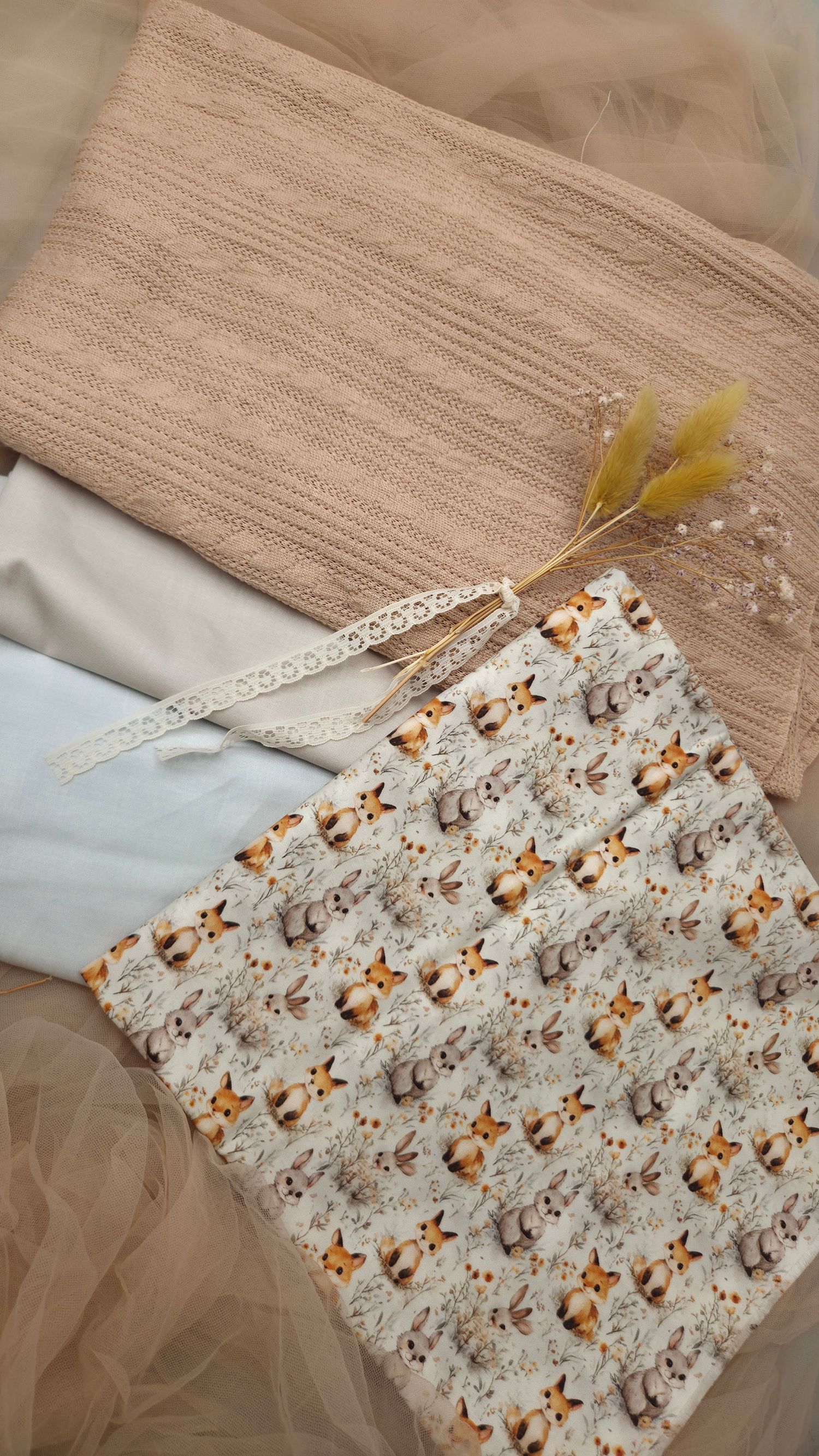 Flat lay of beige knit fabric, light blue cloth, fox print cotton, lace trim, and dried yellow flowers on tulle.
