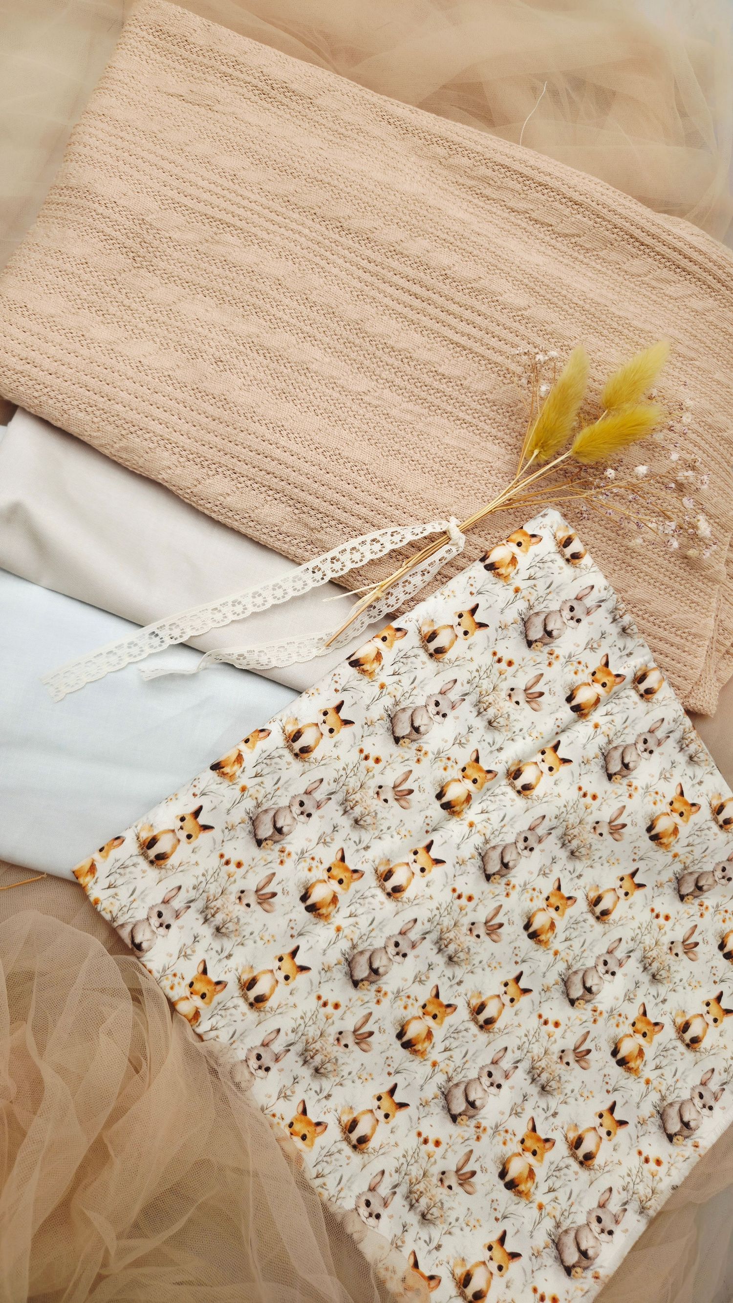 Flat lay of beige knit fabric, white cloth, lace trim, and woodland animal print fabric with dried flowers.
