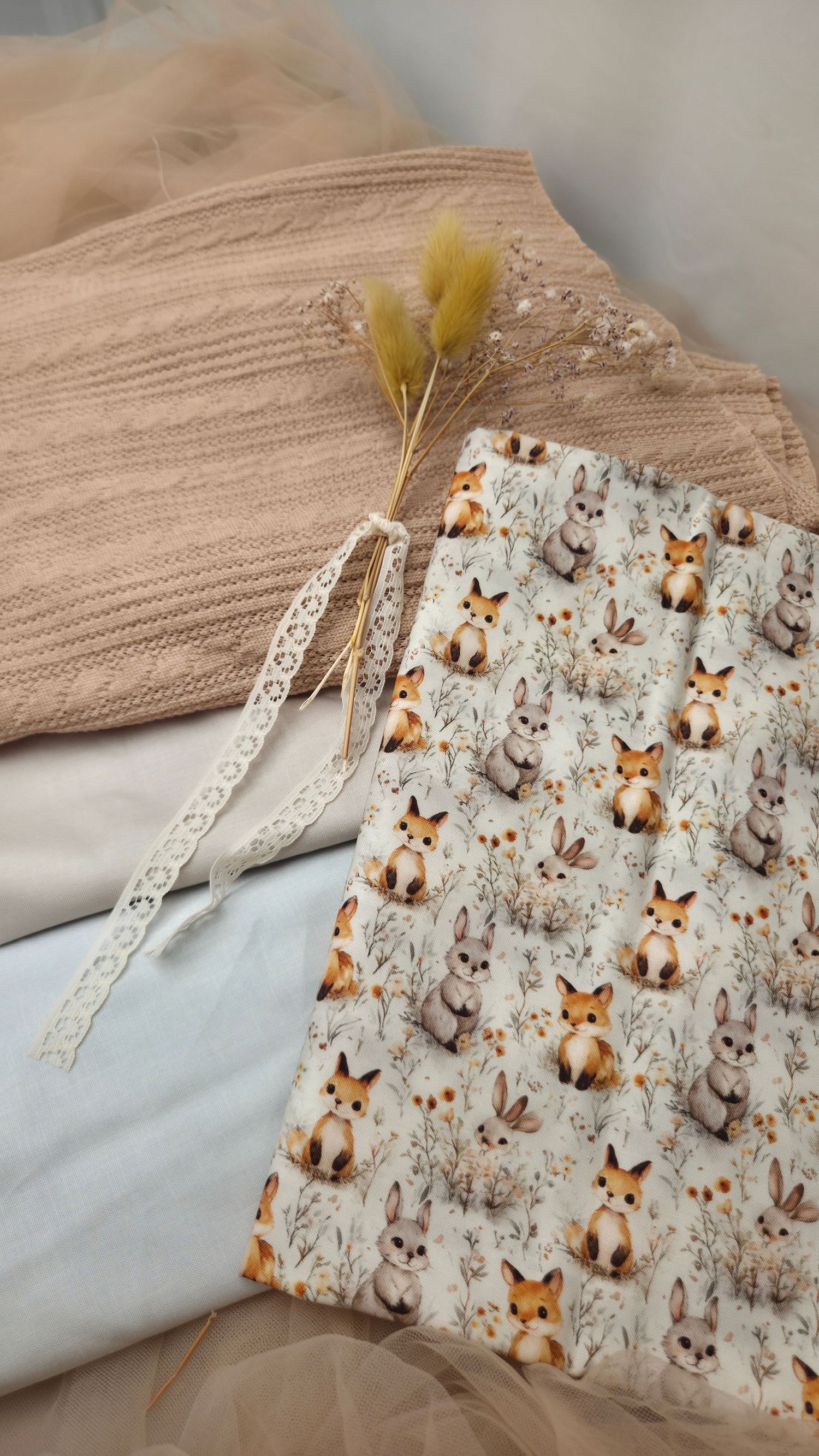 Cute woodland animal fabric with foxes and bunnies beside a beige knit blanket, lace ribbon, and dried flowers.