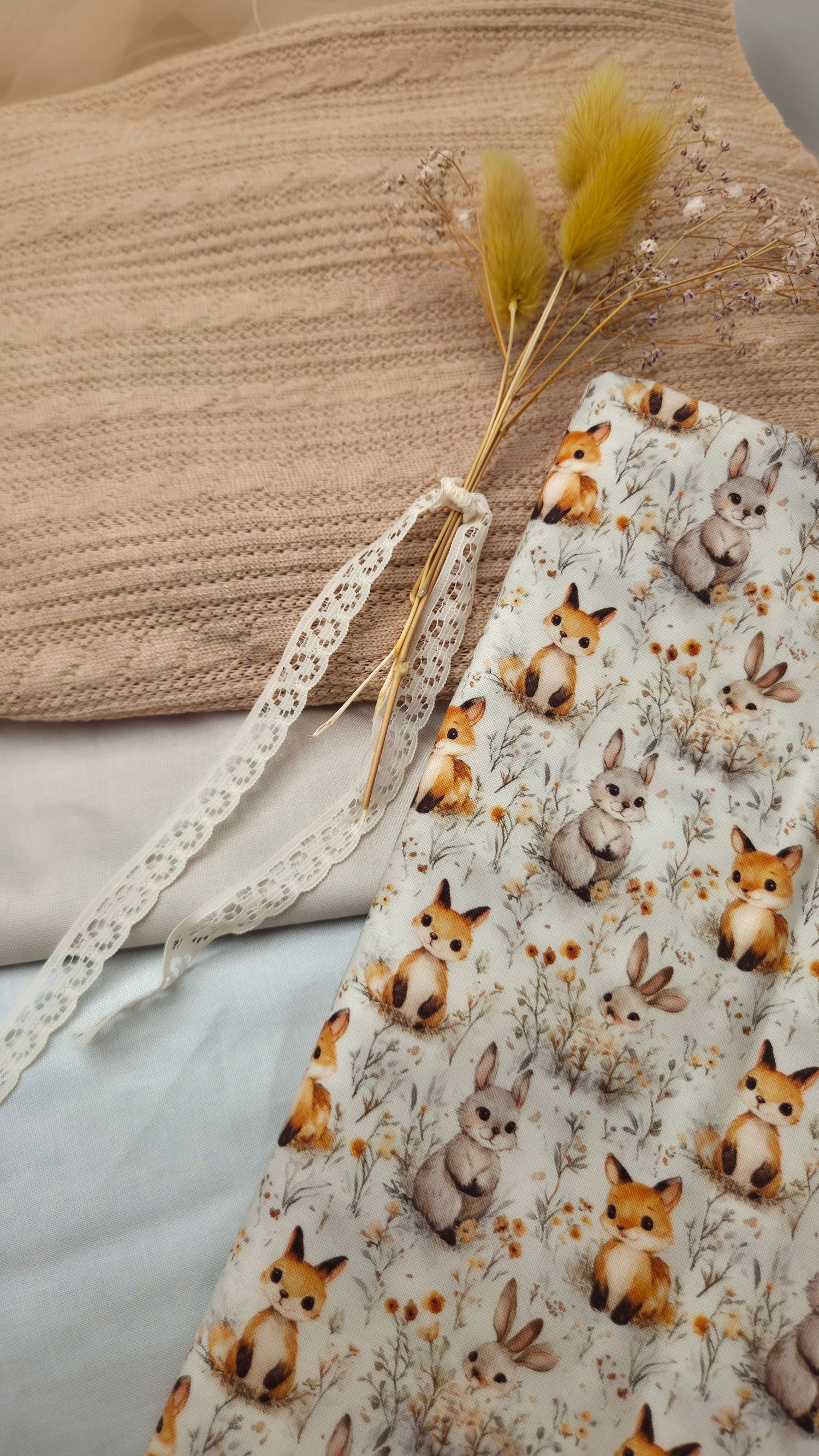 Cozy flat lay featuring fox and bunny patterned fabric, lace trim, dried pampas grass, and a knit pillow in neutral tones.