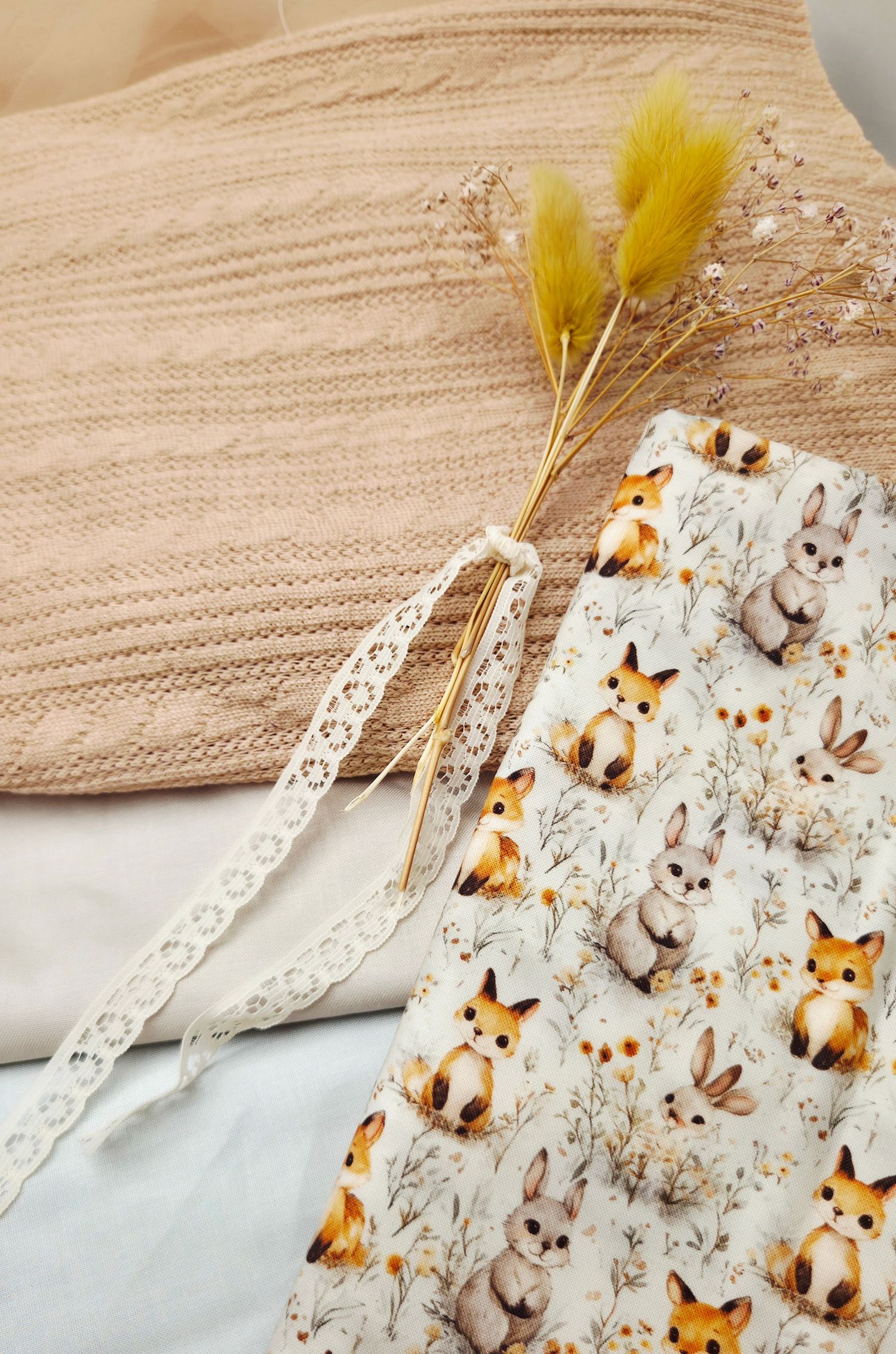 Cozy flat lay with knit pillow, dried flowers, lace ribbon, and woodland animal fabric featuring foxes and bunnies.