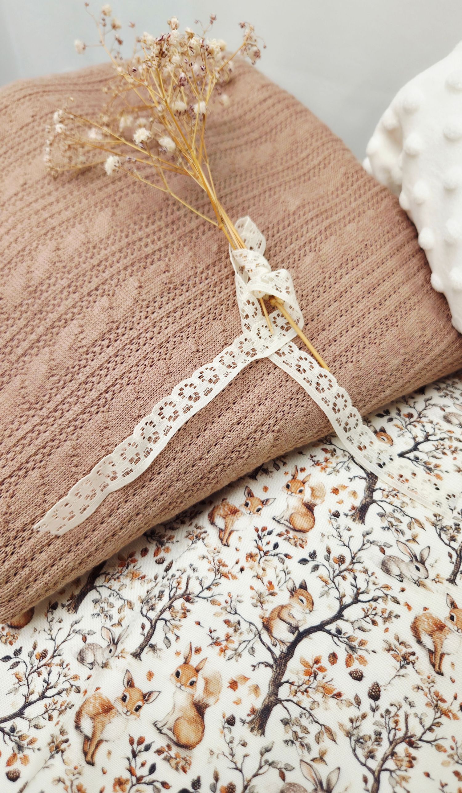 Folded dusty rose knit blanket with lace ribbon and dried flowers on a fox-print fabric background.