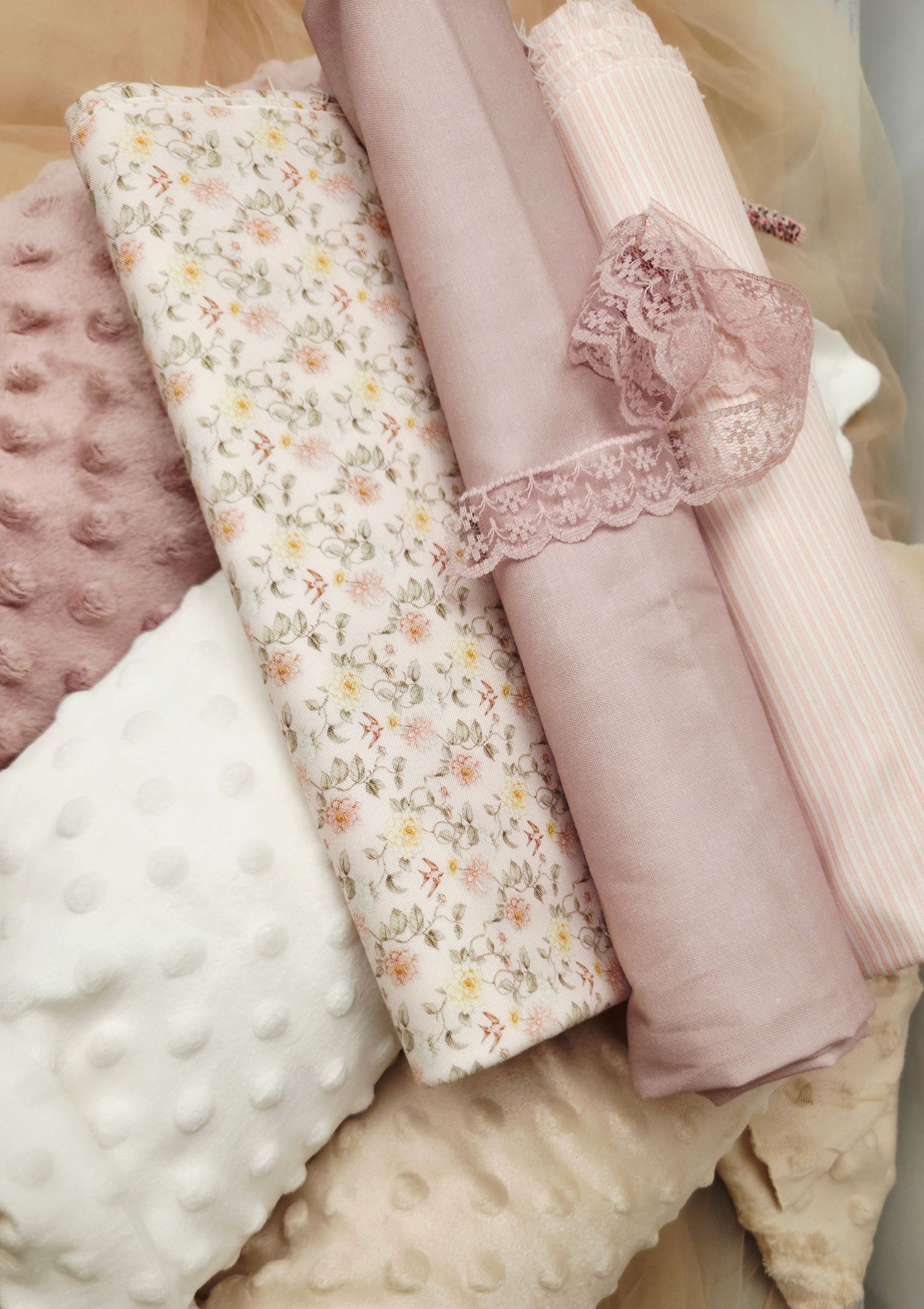 Rolled pink floral and solid fabric swatches with lace trim and textured minky fabric arranged together.