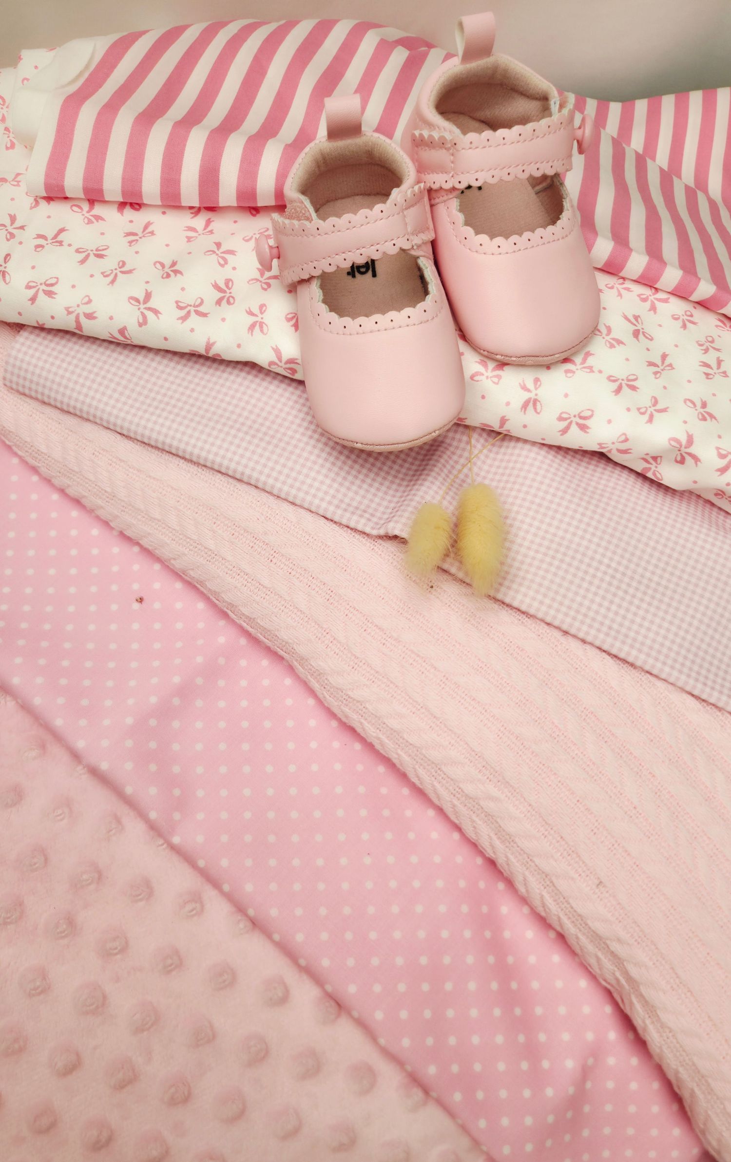 Pink baby girl essentials including soft blankets, floral fabric, striped pillow, and tiny pink leather Mary Jane shoes.