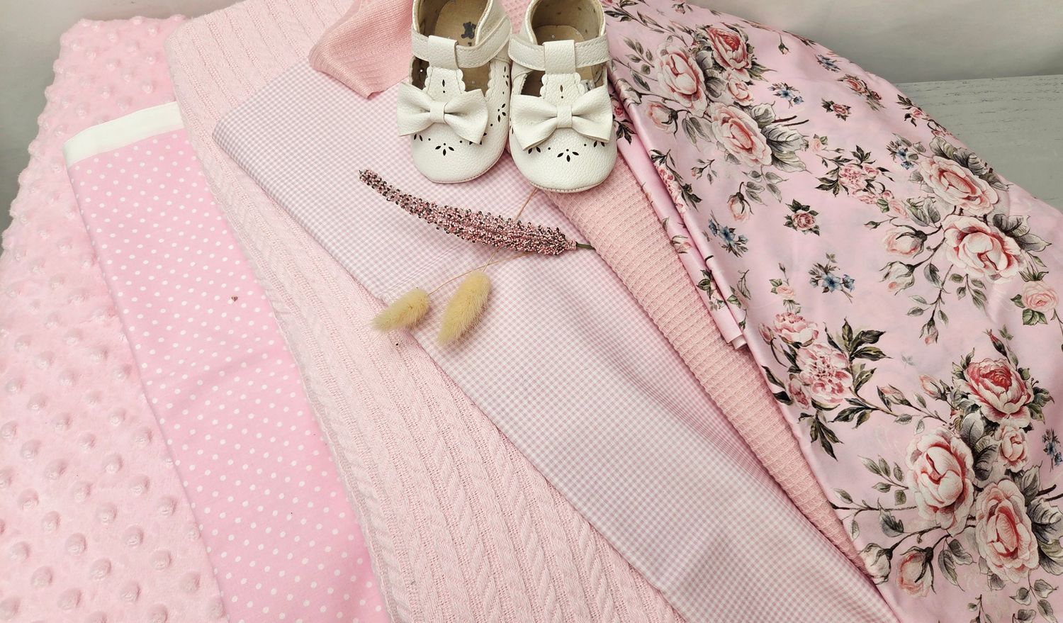 Pink baby girl layette set with floral fabric, polka dot blanket, striped cloth, feathers, and white bow shoes.