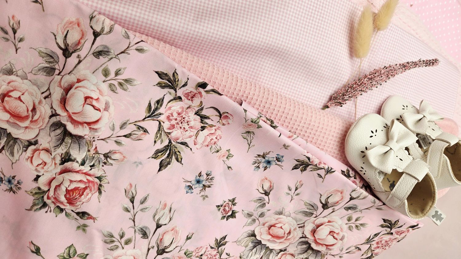 Pink floral fabric with roses, pink gingham textile, and white baby shoes arranged on a soft pink background.