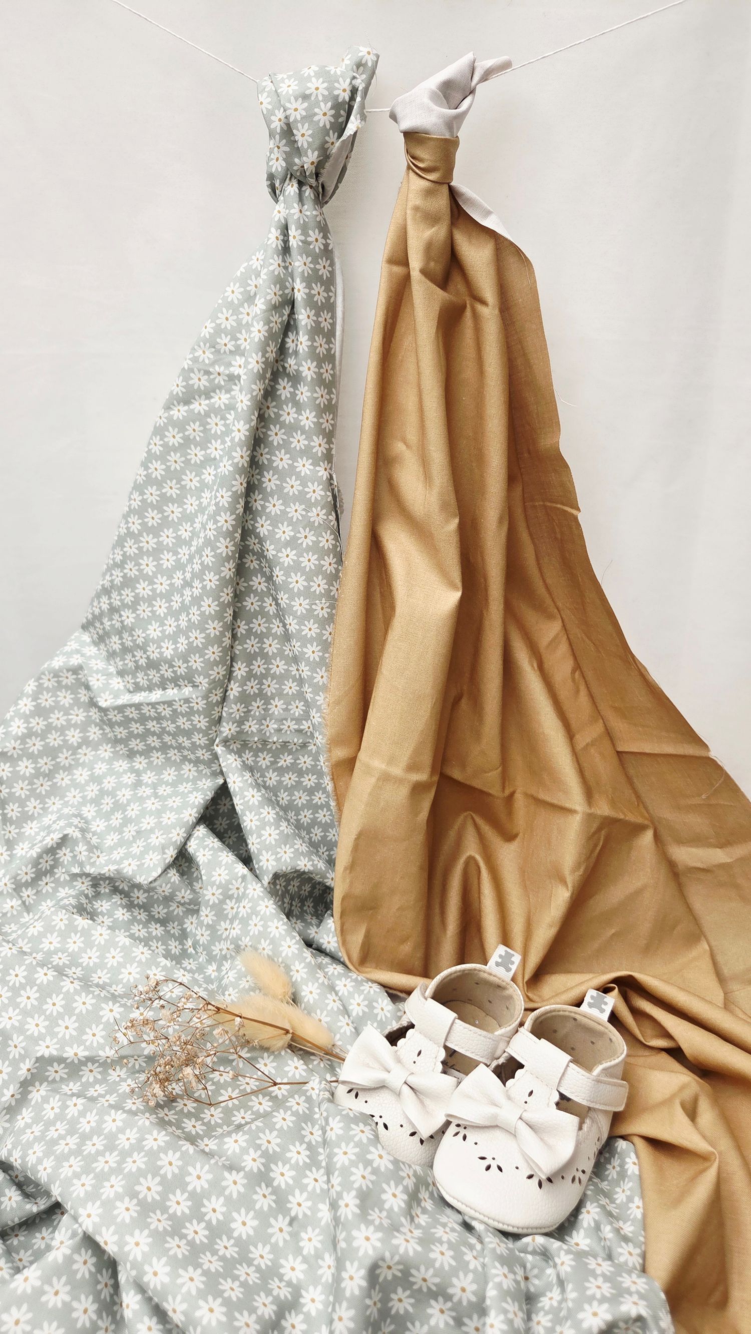Two swaddle blankets in tan and floral grey hang draped with white baby shoes and dried flowers below.