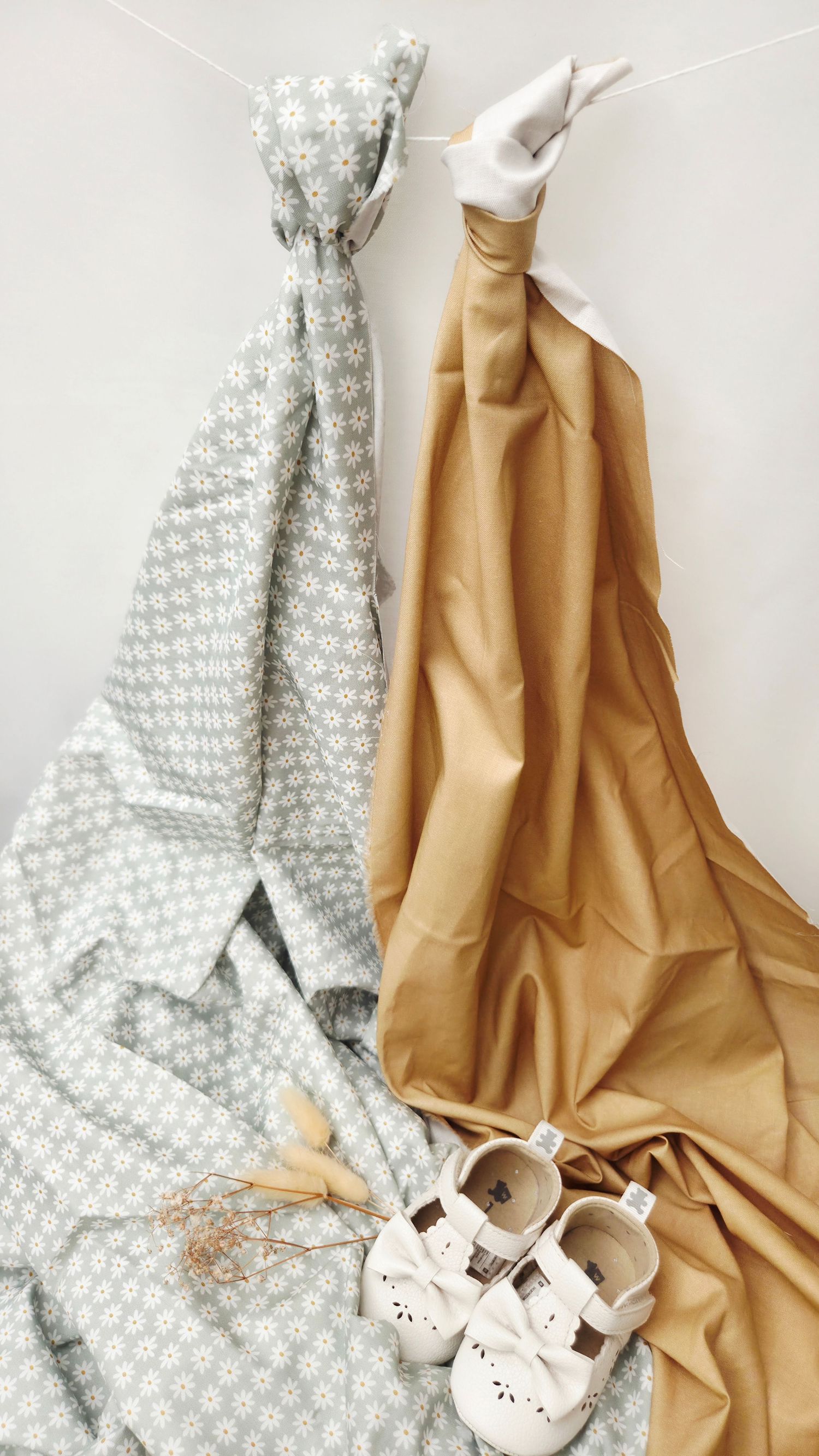 Two baby swaddle blankets in gray floral and tan hanging with white infant shoes and dried flowers below.
