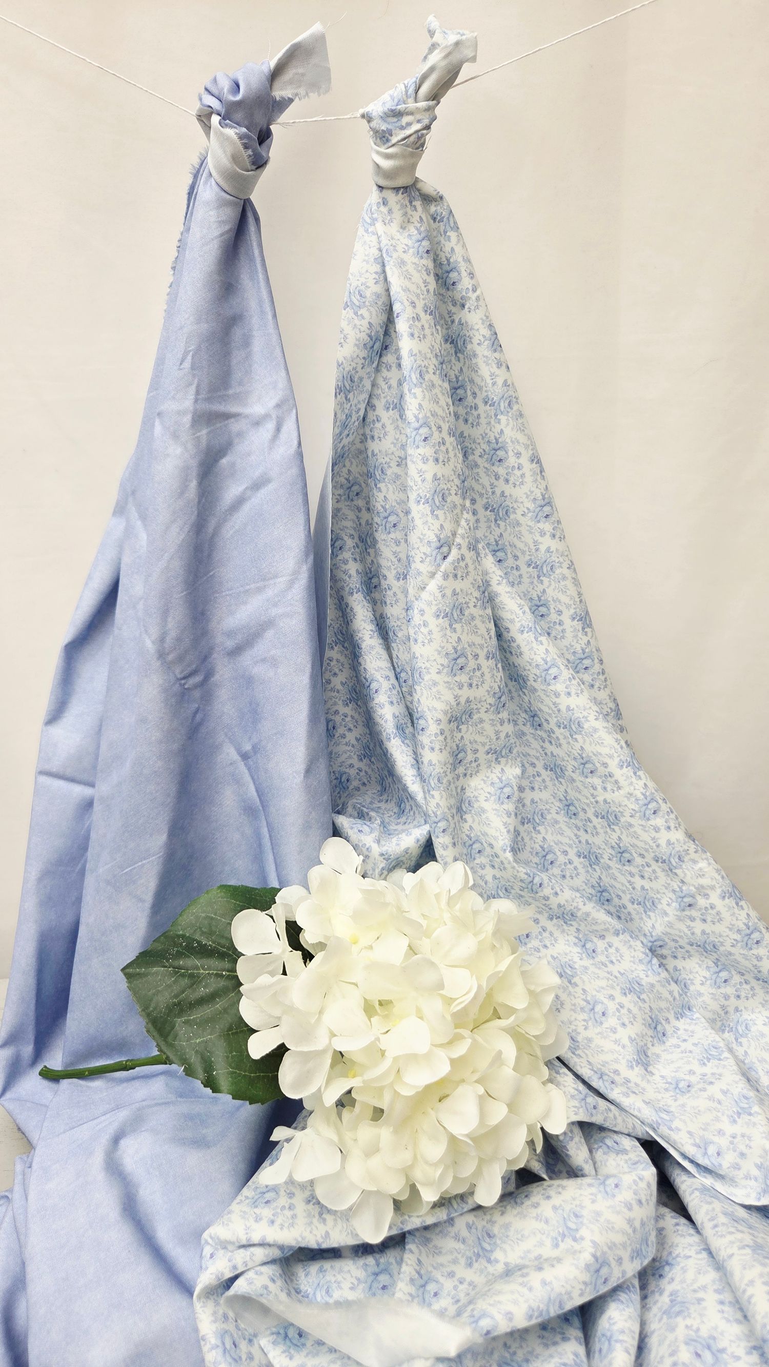 Two blue swaddle blankets, one solid and one floral, draped with a white hydrangea flower in front.