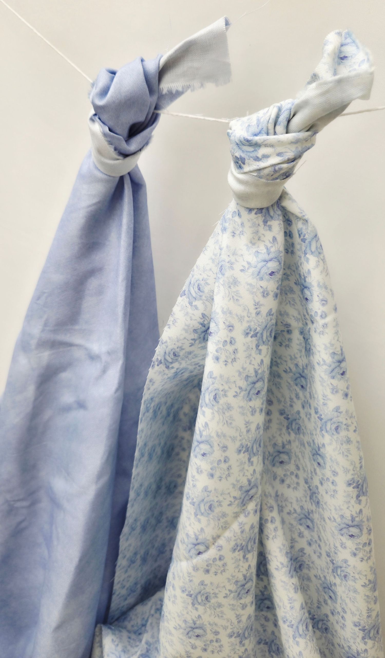 Two blue fabric swaddle blankets, one solid and one floral, tied to a clothesline against a white wall.