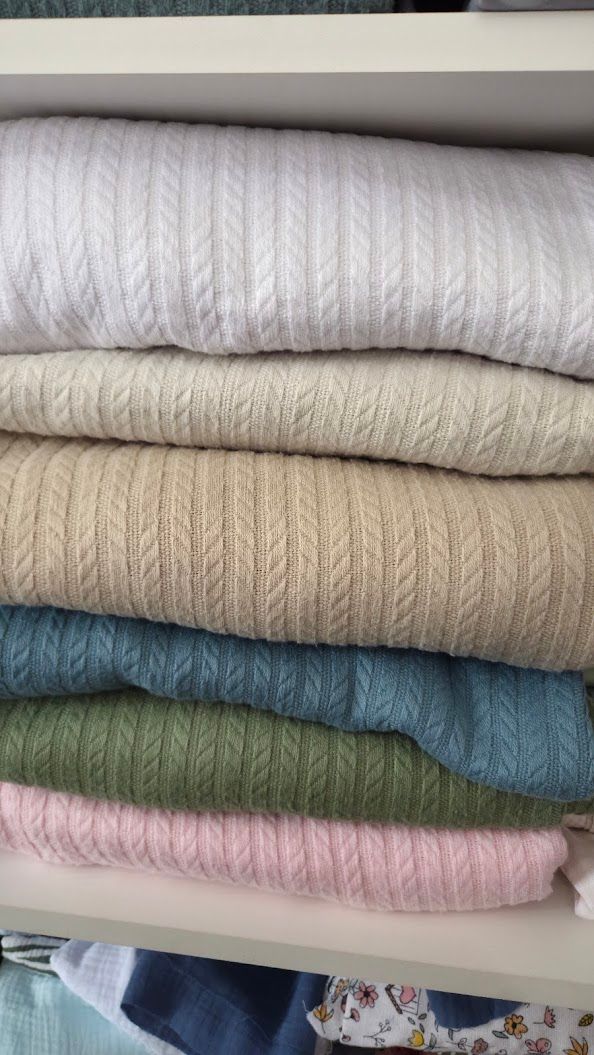 Stack of neatly folded cable-knit sweaters in white, beige, tan, teal, green, and pink on a white shelf.