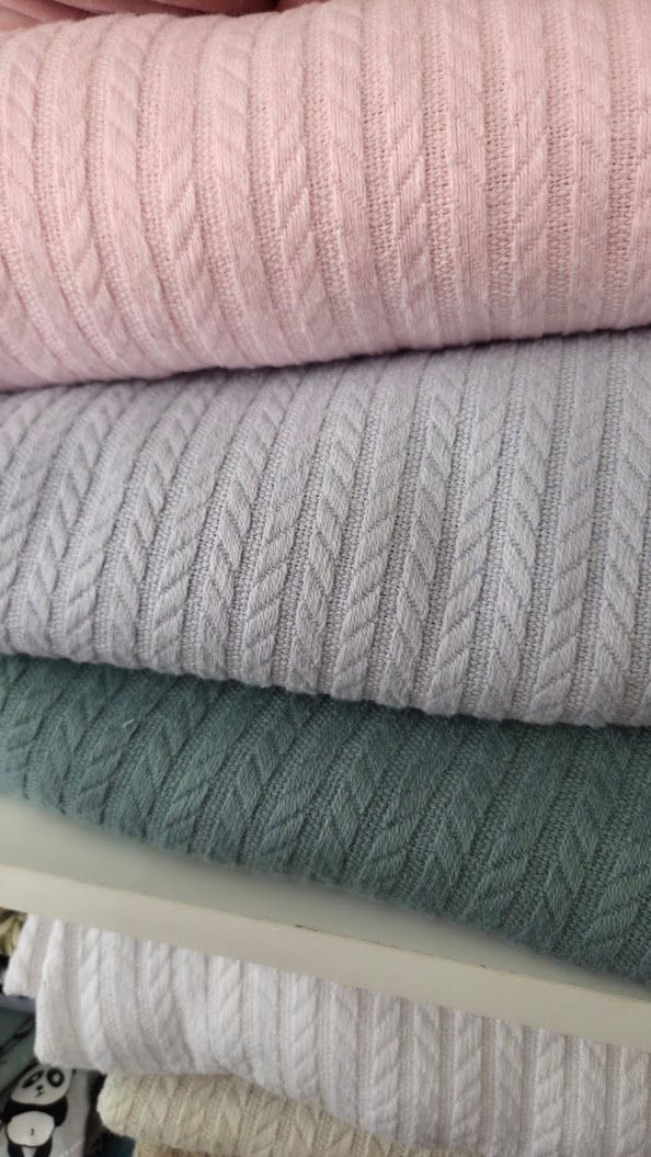 Stack of folded cable-knit sweaters in pink, lavender, and teal pastel colors neatly arranged on a shelf.