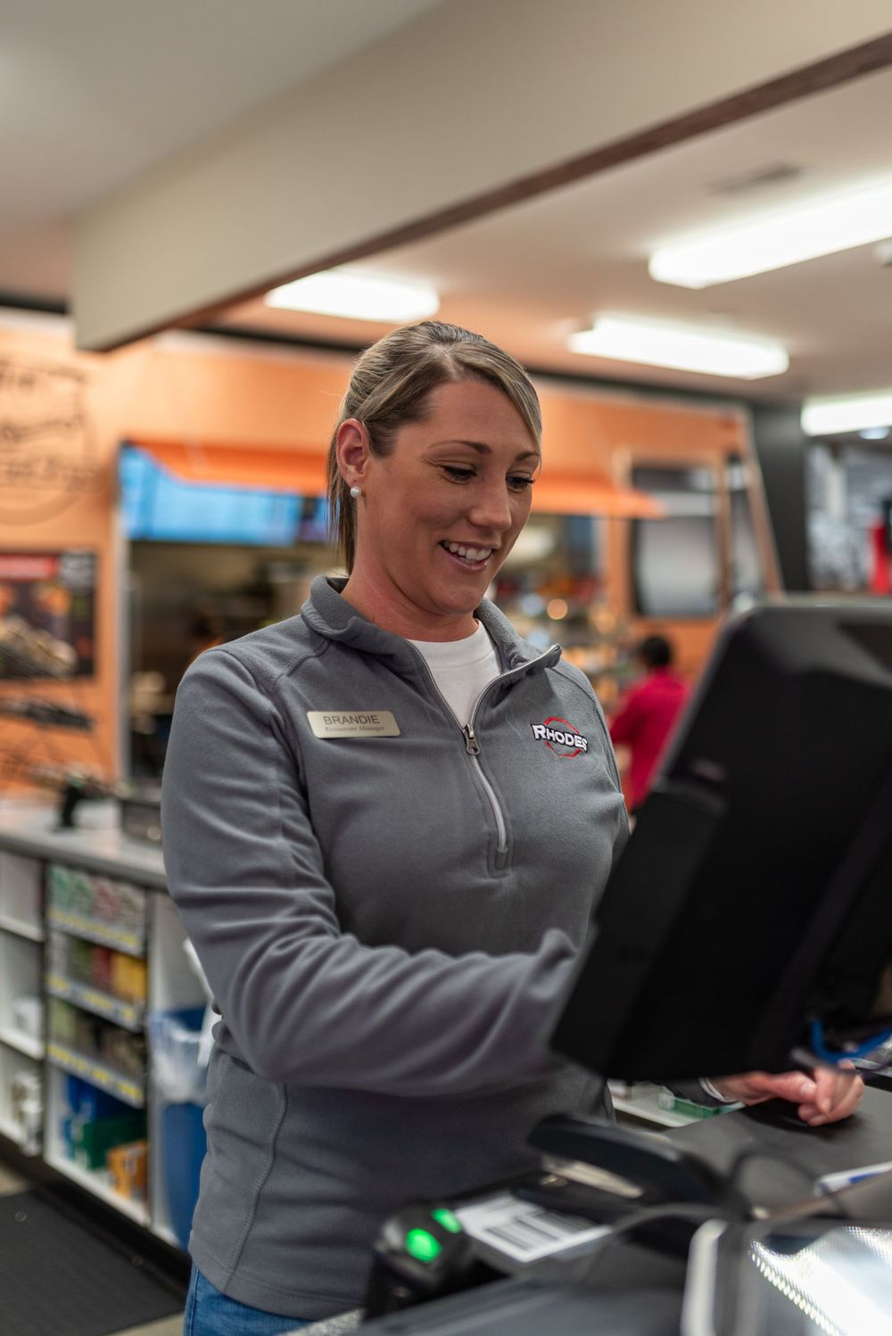 Smiling female cashier in gray uniform jacket working at a retail store checkout counter with a touchscreen POS system.
