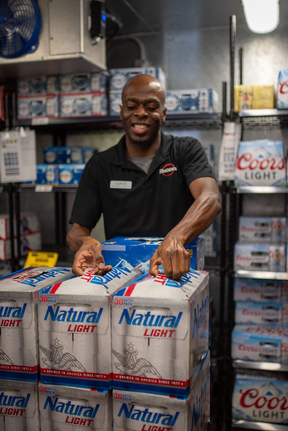 Smiling store employee stacking Natural Light beer cases in a beverage storage room with Coors Light visible.