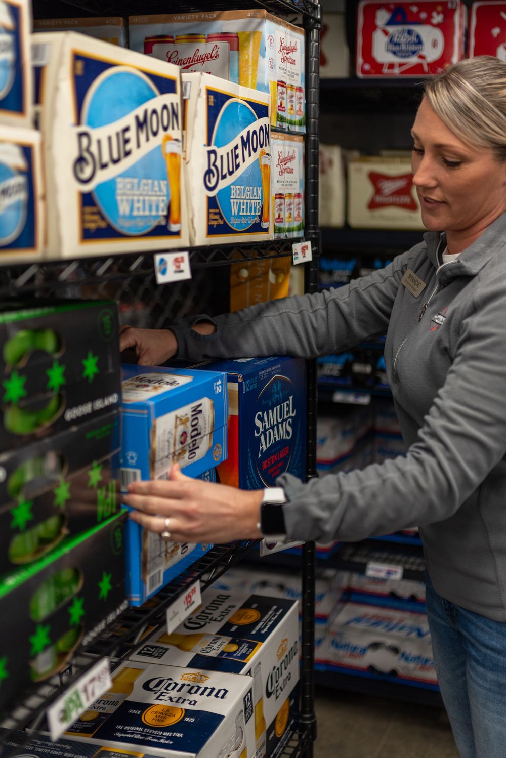 Woman stocking shelves with Blue Moon, Samuel Adams, and Corona beer cases in a retail store.