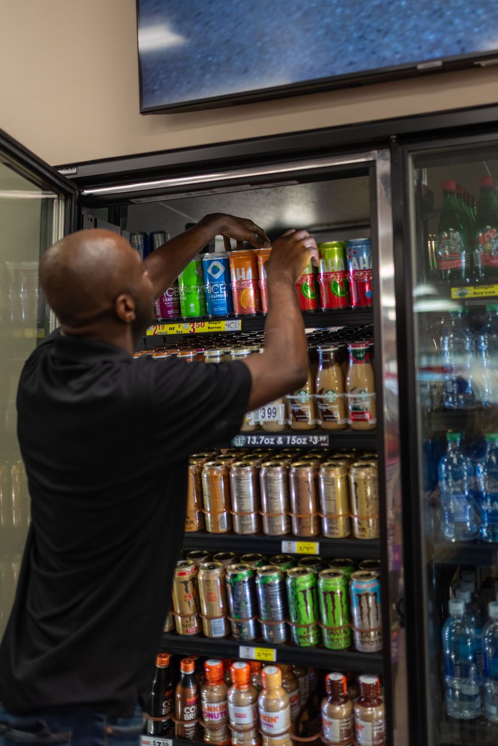 Man restocking colorful energy drinks and beverages in a commercial refrigerator display case at a convenience store.