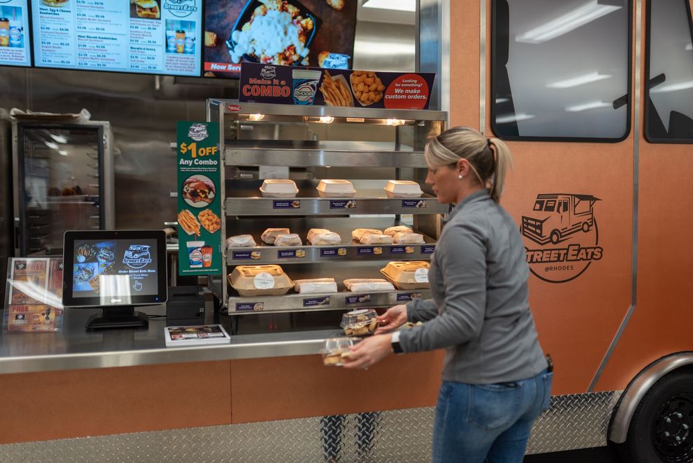 Woman ordering food at an orange food truck window with displayed menu items and baked goods on shelves.
