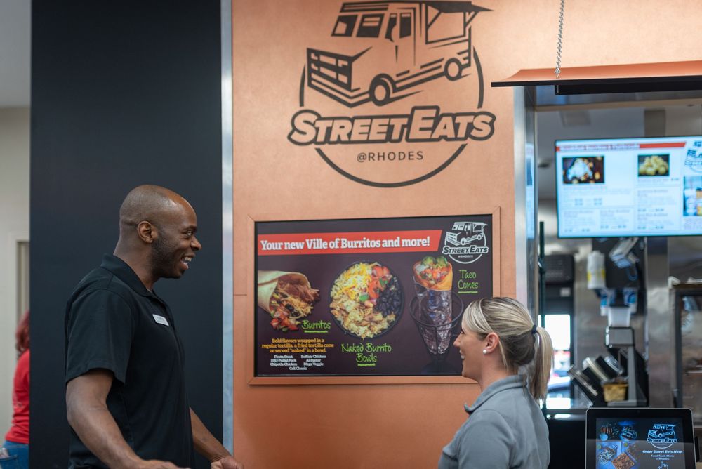 Two people conversing at Street Eats by Rhodes food counter with colorful burrito menu display on wall.