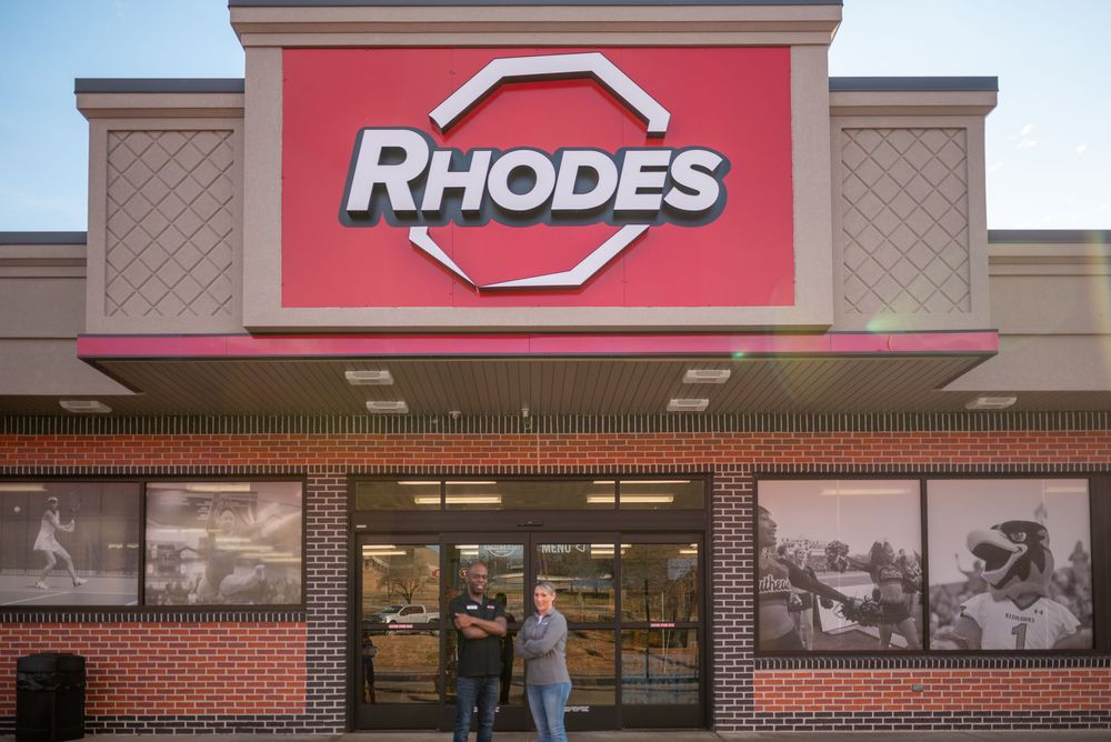 Two employees stand outside a Rhodes convenience store with brick facade and bold red signage.