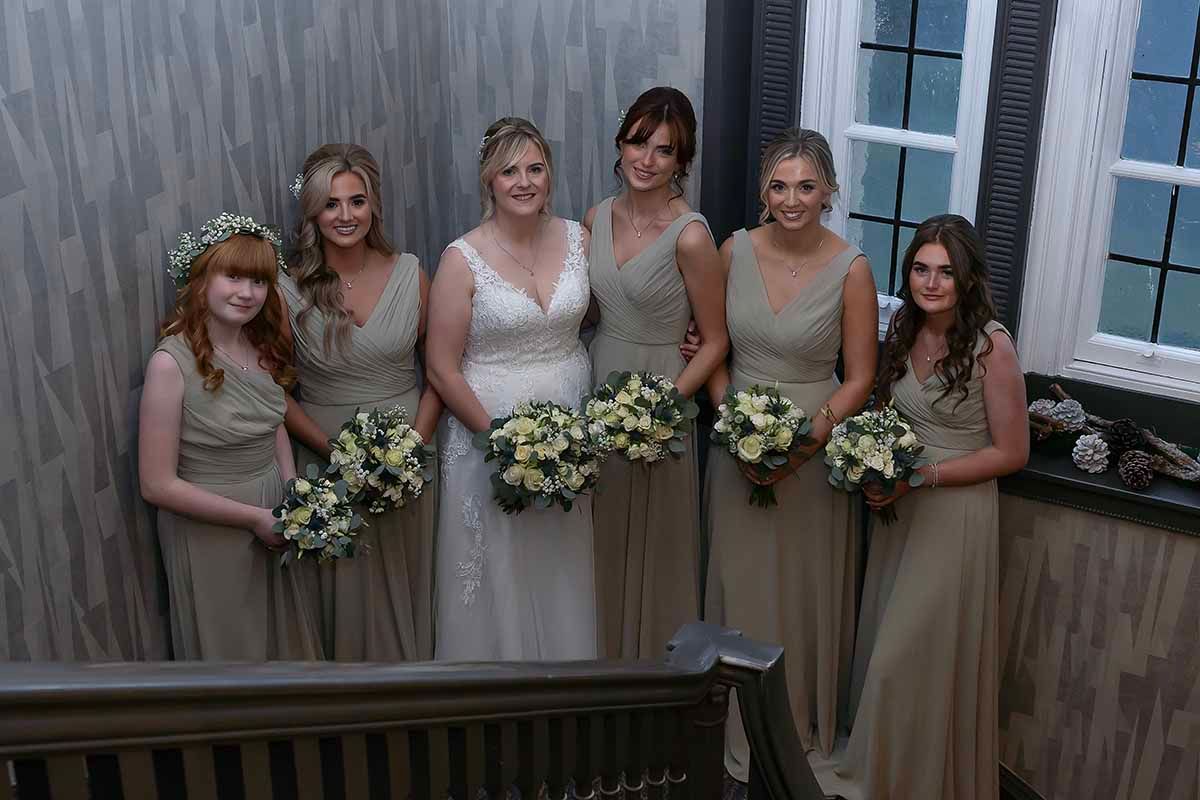 Bride in white gown poses with four bridesmaids in champagne dresses holding bouquets on the grand staircase at Hetland Hall