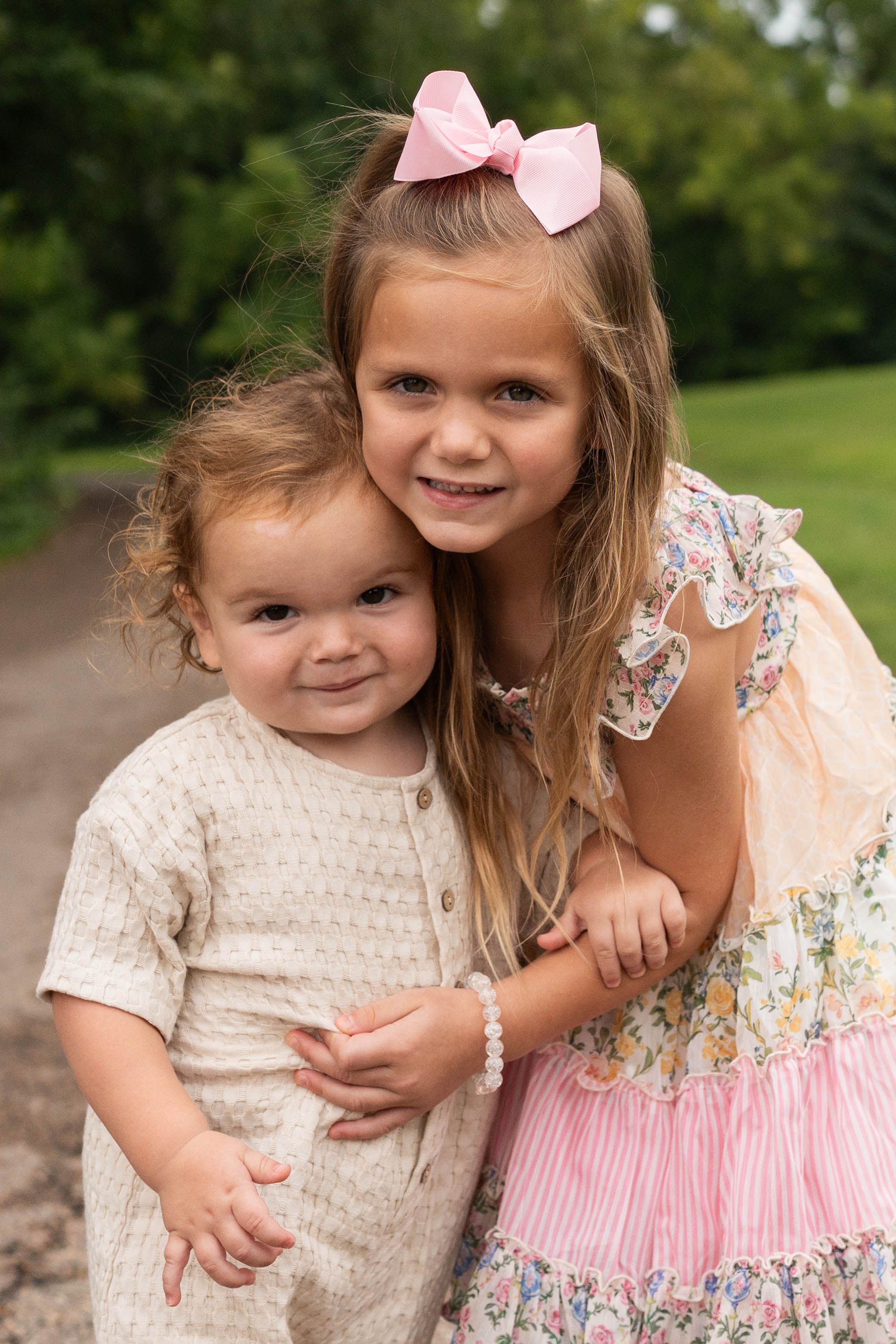 Two young sisters pose outdoors, older girl in floral dress with pink bow hugging toddler in cream romper.