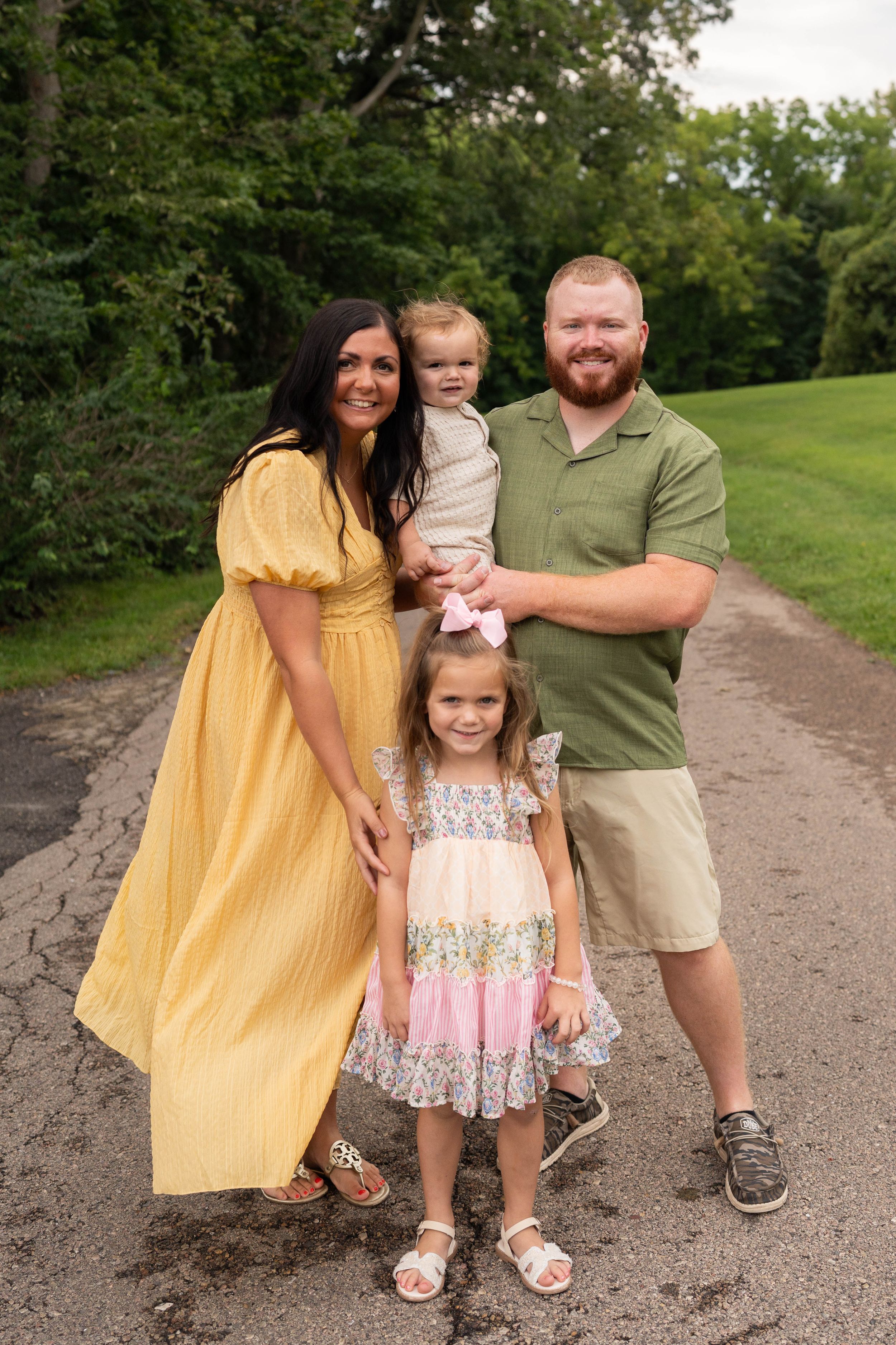 Happy family of four posing on a tree-lined path, mom in yellow dress, dad in green shirt holding toddler.