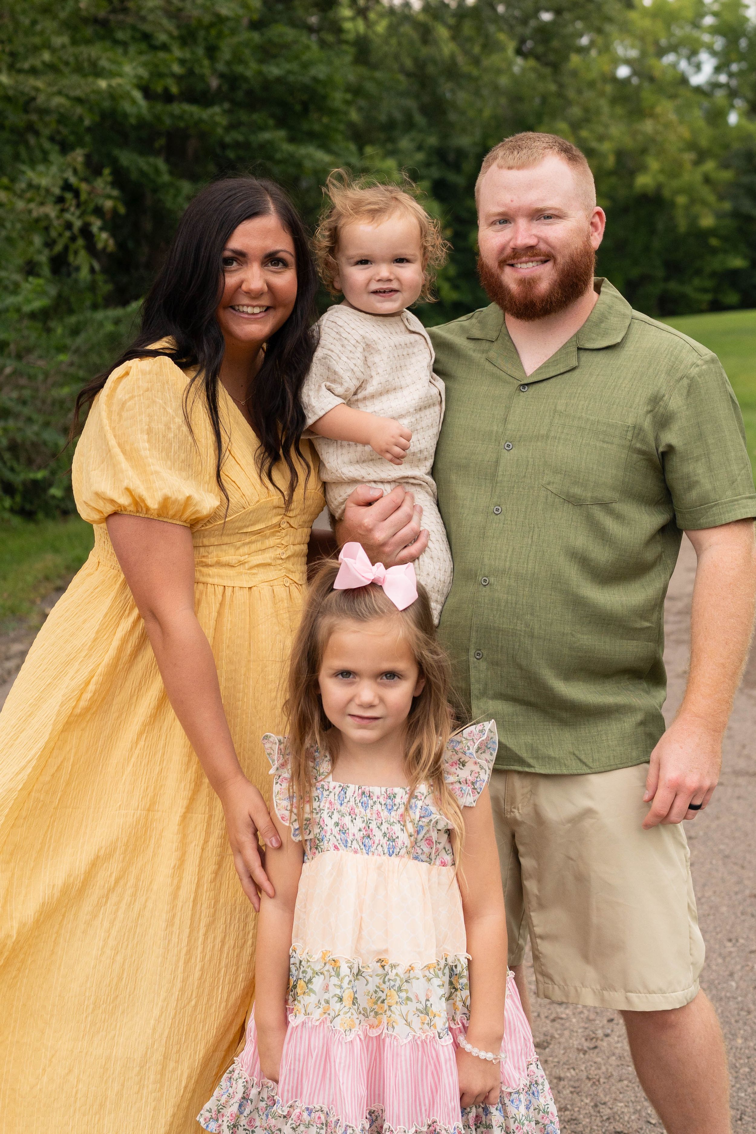 A smiling family of four poses outdoors, mom in yellow dress, dad in green shirt, holding two young daughters.