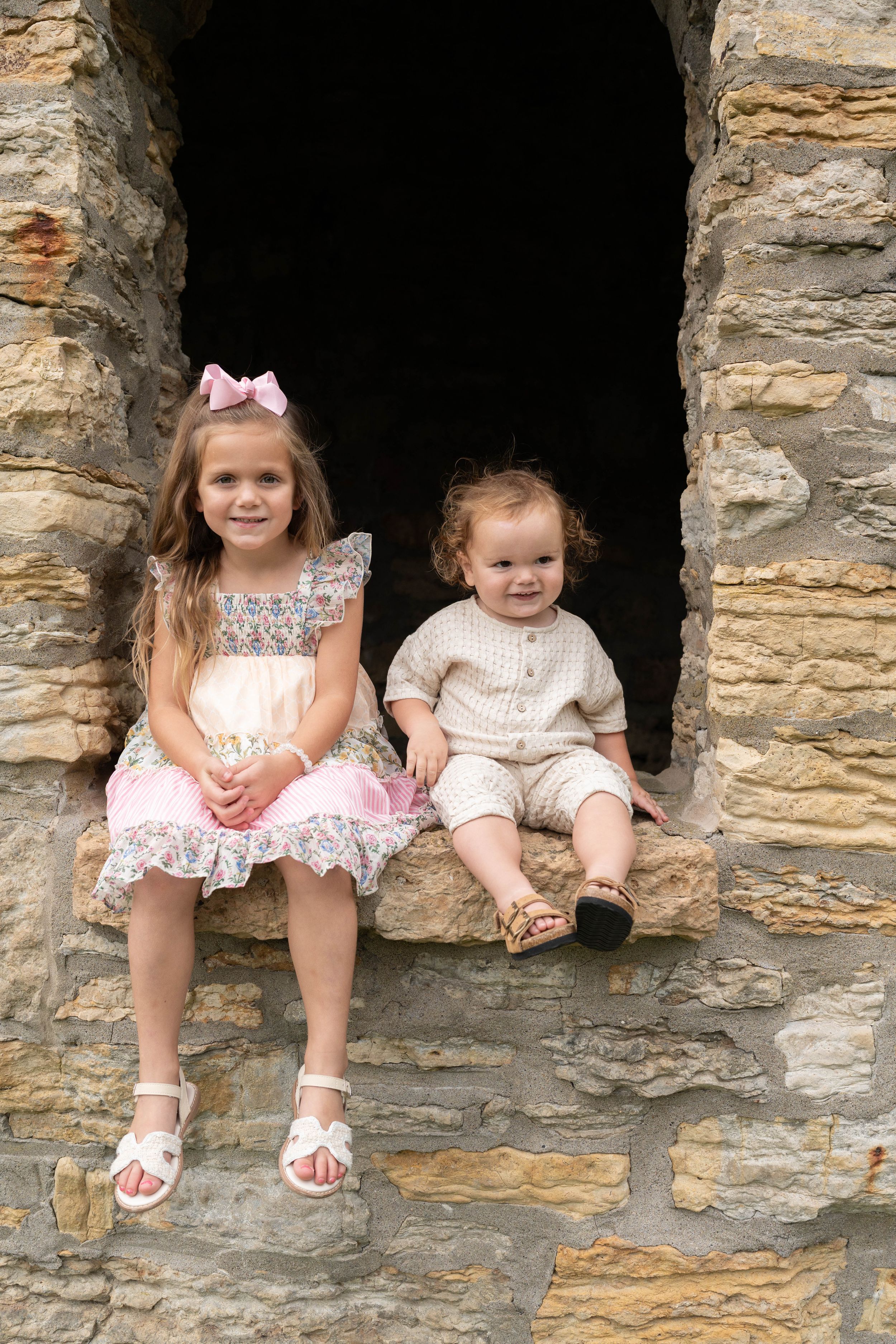 Two young children sitting in a rustic stone window opening, girl in floral dress with pink bow, toddler in beige outfit.