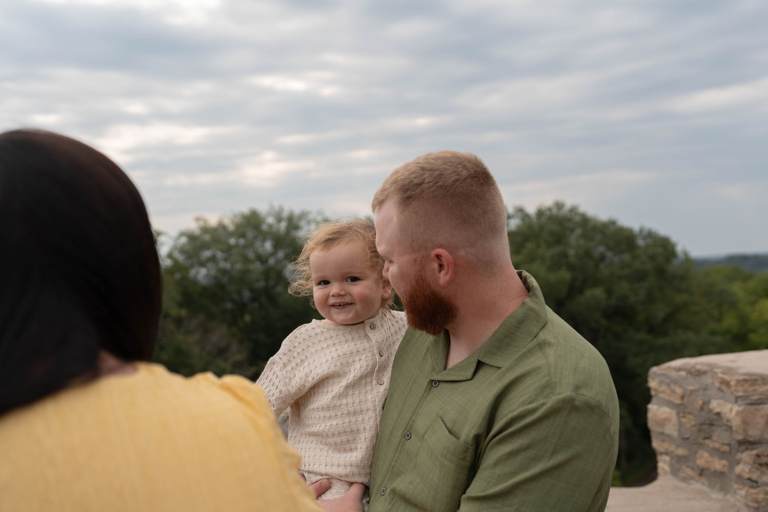 A man in a green shirt holds a smiling baby while a woman in yellow stands nearby outdoors.