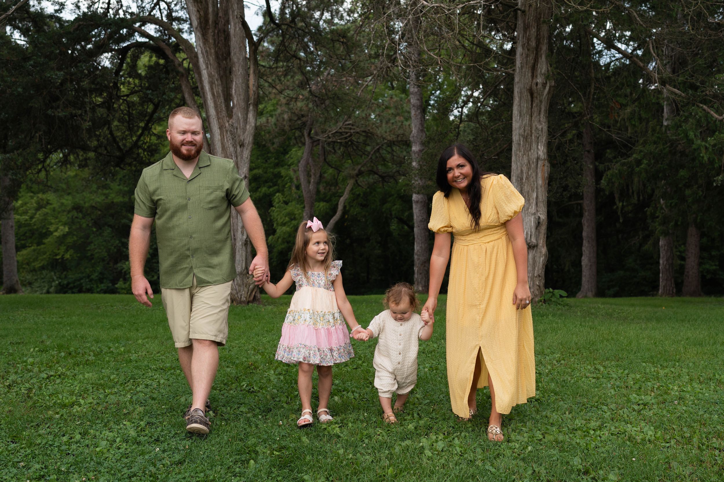 Family of four holding hands and walking on green grass surrounded by tall trees in a park setting.