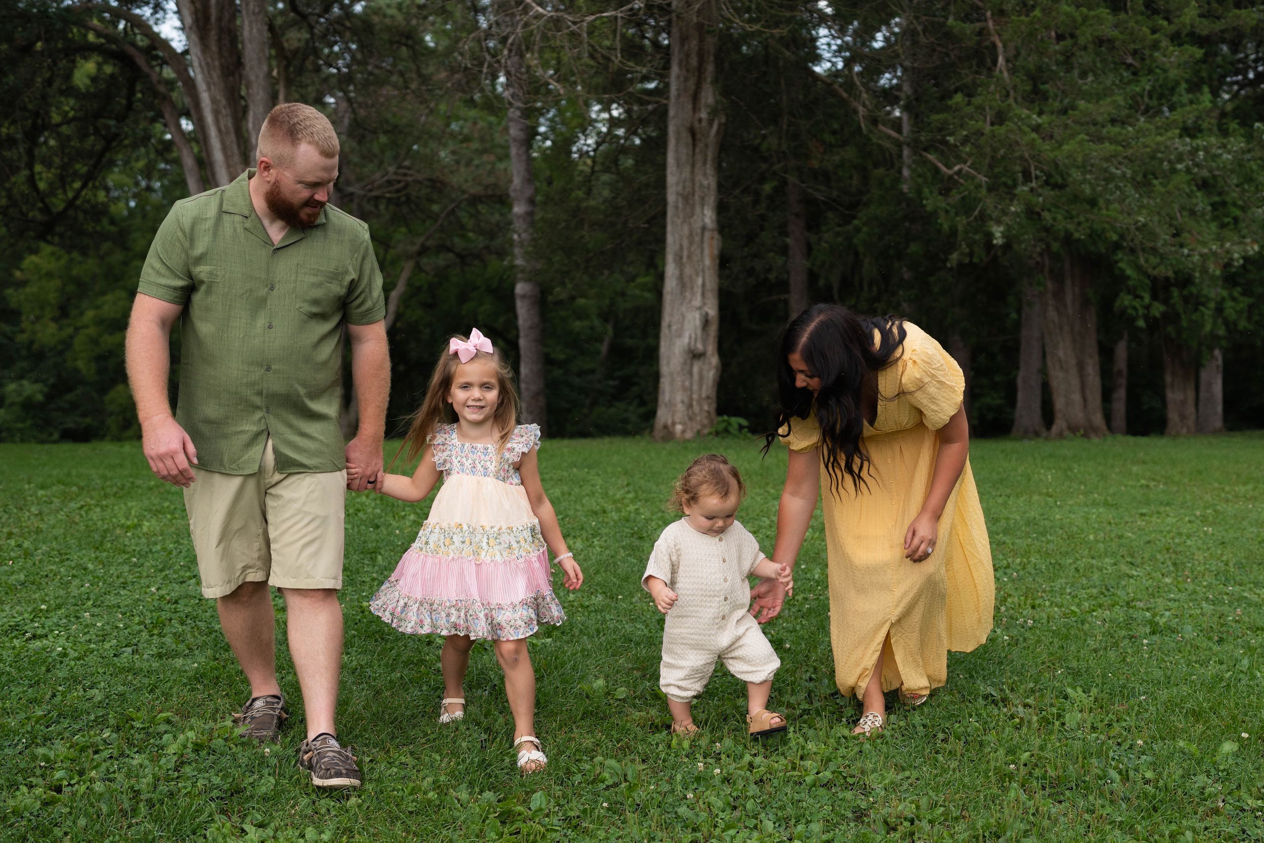 Family of four walking on green grass in a park, parents holding hands with young children in summer outfits.