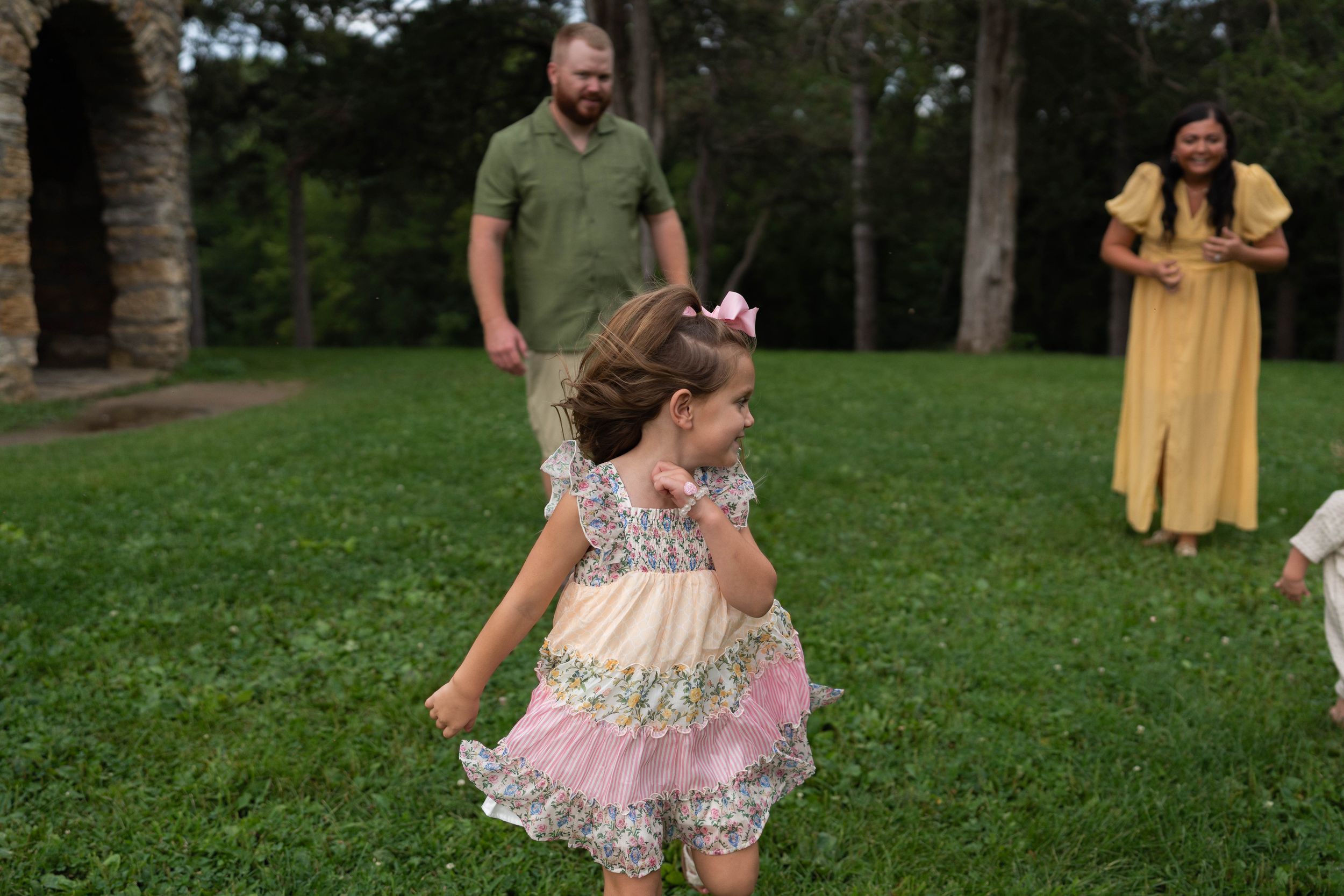 A young girl in a pink floral dress runs on green grass while her parents watch and smile outdoors.