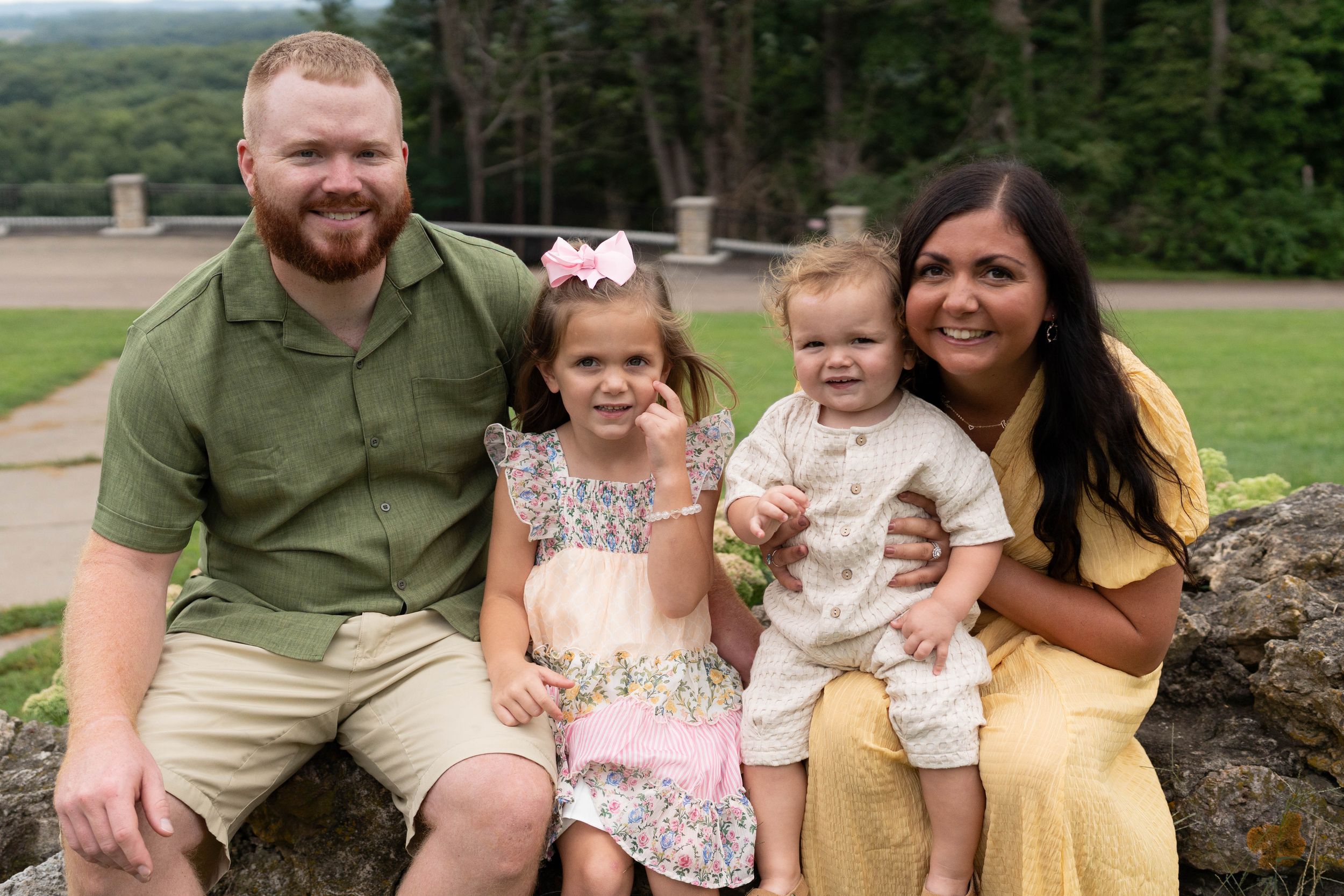 A happy family of four poses outdoors, parents smiling with two young children in coordinated outfits.