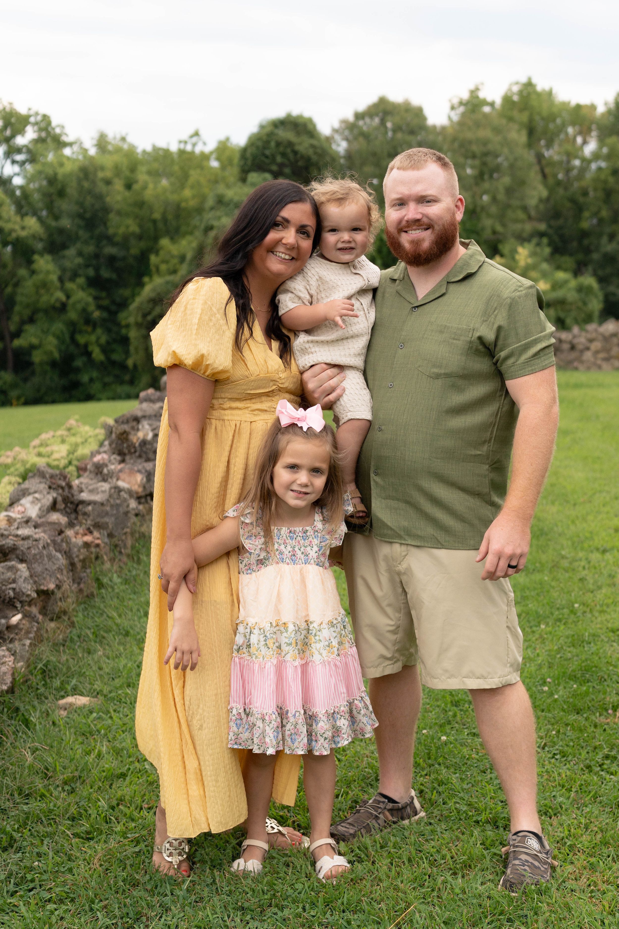 A happy family of four poses outdoors on green grass, with mom in yellow dress, dad in green shirt, and two young children.