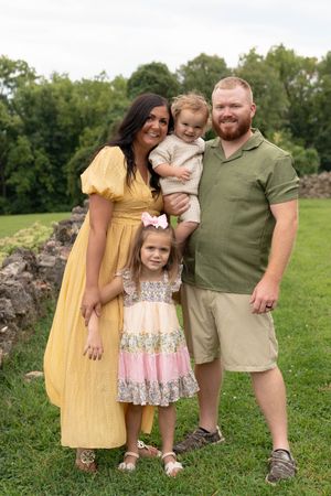 A smiling family of four poses outdoors on green grass, with a woman in yellow, man in green, and two young children.