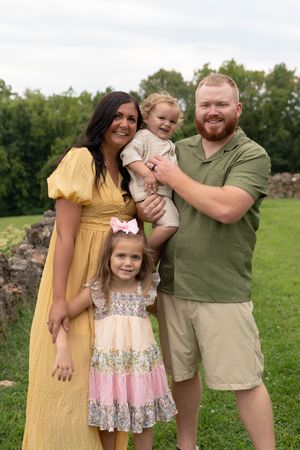 A smiling family of four poses outdoors on green grass, with two parents holding young daughters in colorful summer outfits.