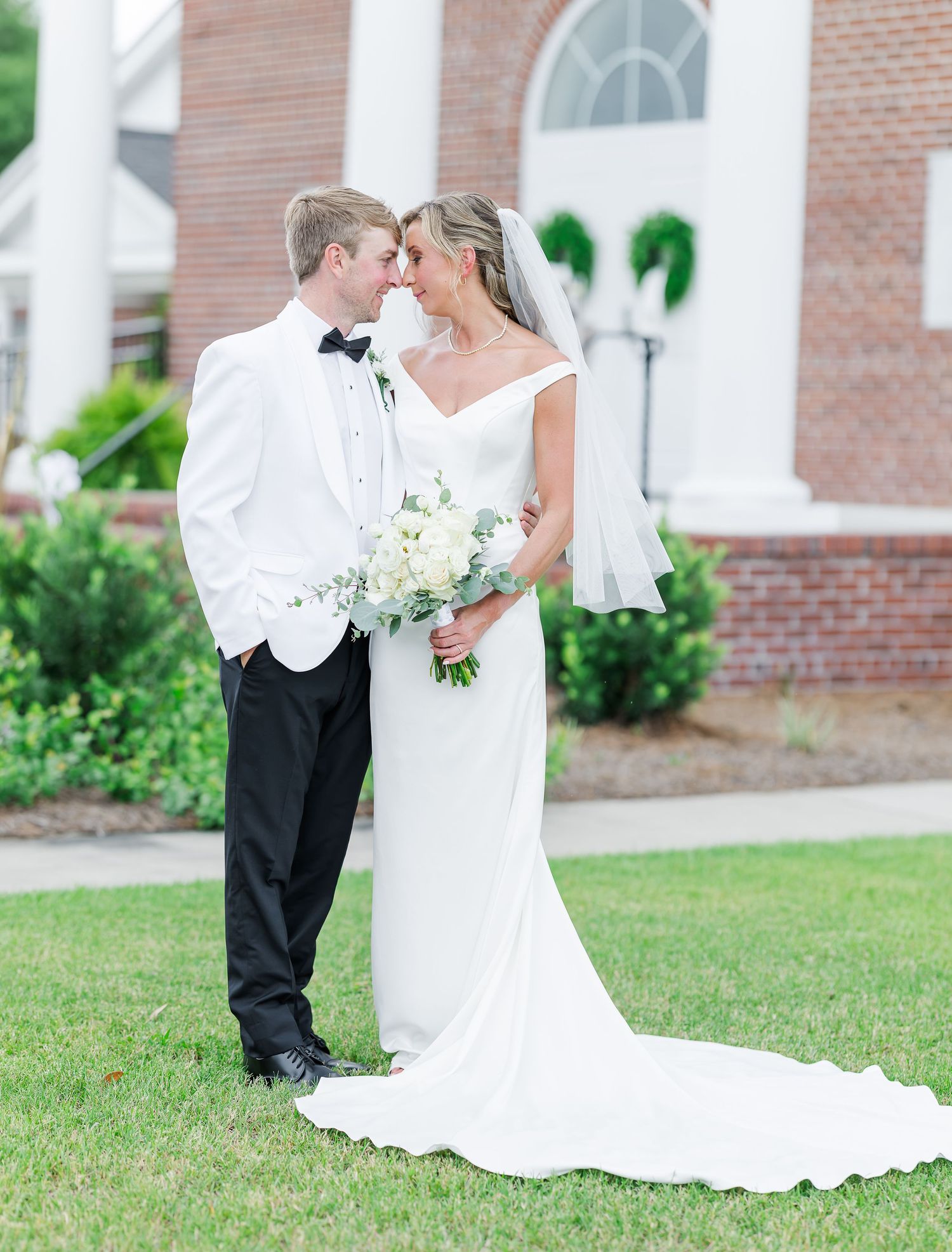 Bride and groom pose on brick steps before white arched door with greenery wreaths at elegant wedding venue.