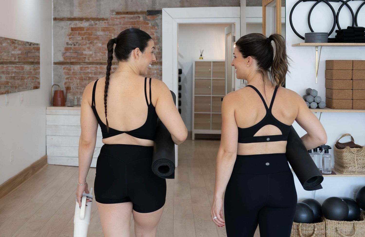 Two women in black activewear holding yoga mats in a fitness studio, viewed from behind.