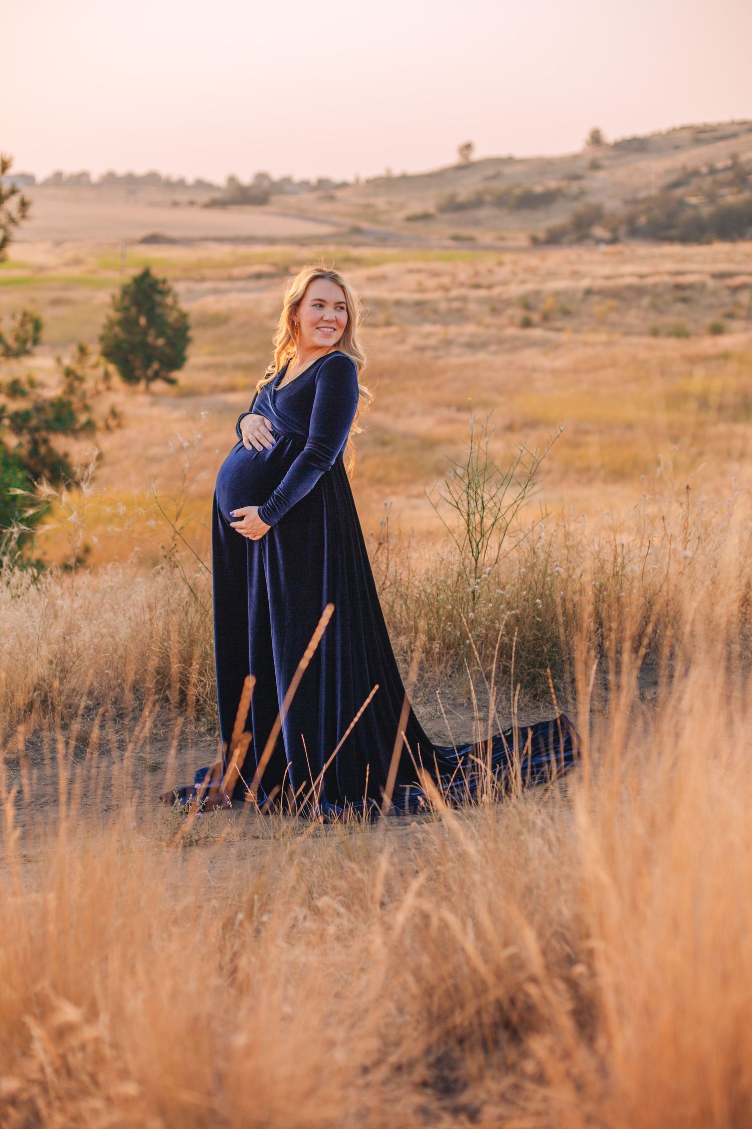 Pregnant woman in navy blue velvet gown standing in golden field at sunset, smiling and cradling her baby bump.